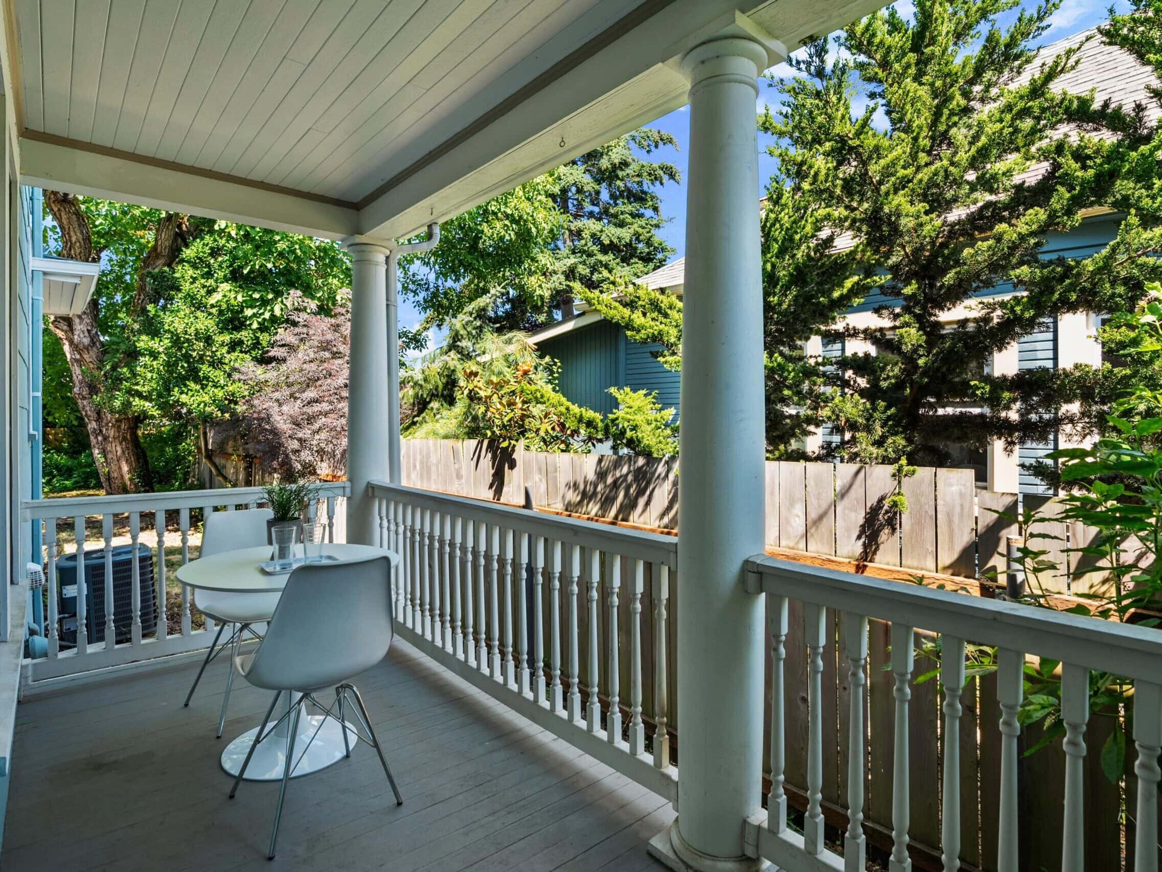 A cozy porch with white railings and columns, featuring a small round table and two white chairs, overlooking green trees and neighboring houses on a sunny day.