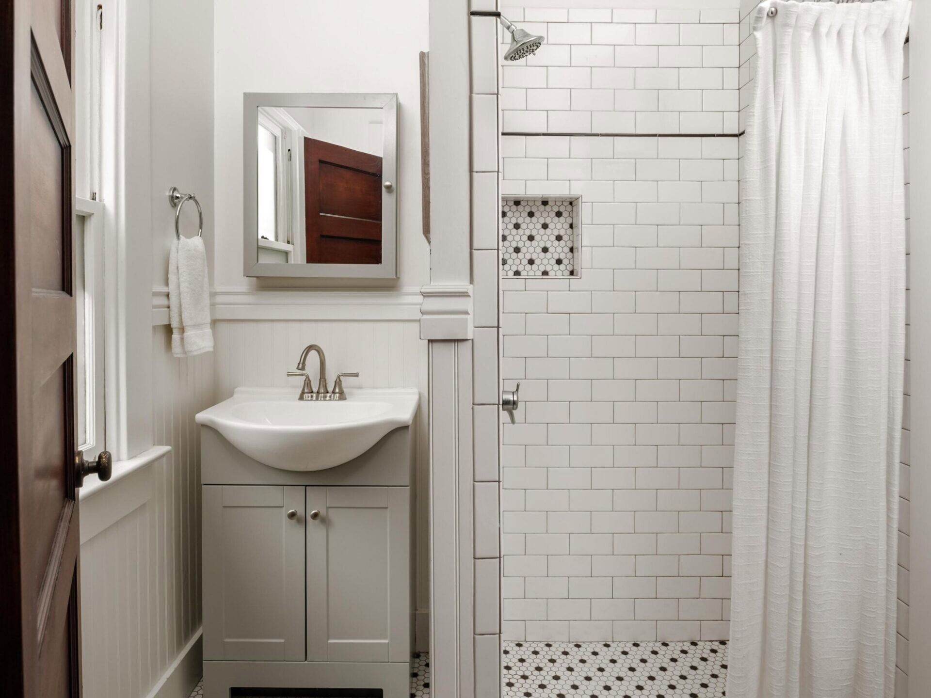 Small white bathroom with hexagonal floor tiles, a pedestal sink with a cabinet, wall mirror, towel ring, and a walk-in shower with white subway tiles and a shower curtain.