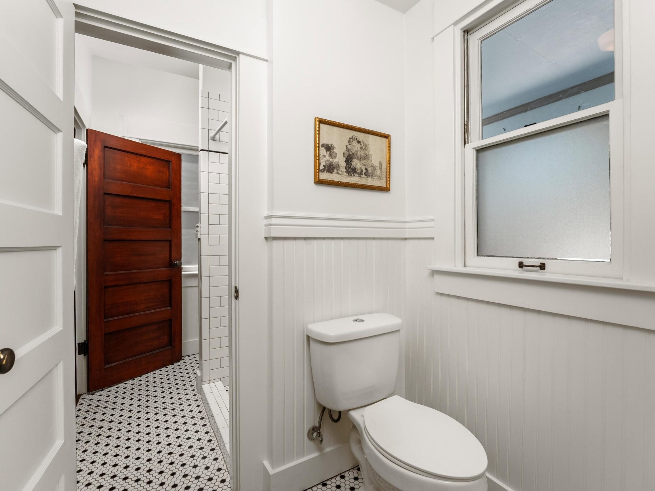 A small white bathroom with a toilet, a window with frosted glass, a framed picture above the toilet, and a view into a shower area with white tiles and a wooden door.