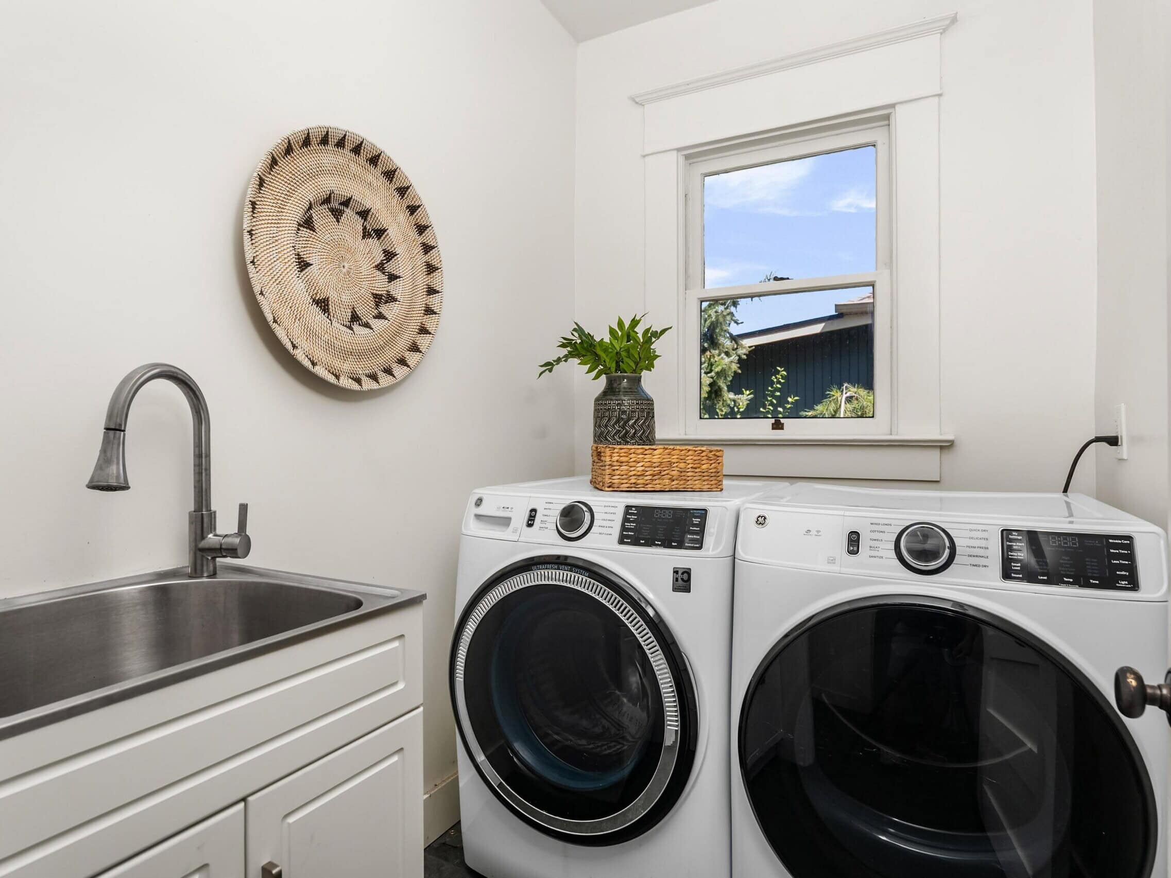 A modern laundry room with a white sink, washer, and dryer. A woven wall decoration hangs above, and a plant in a vase sits on a basket by the window.