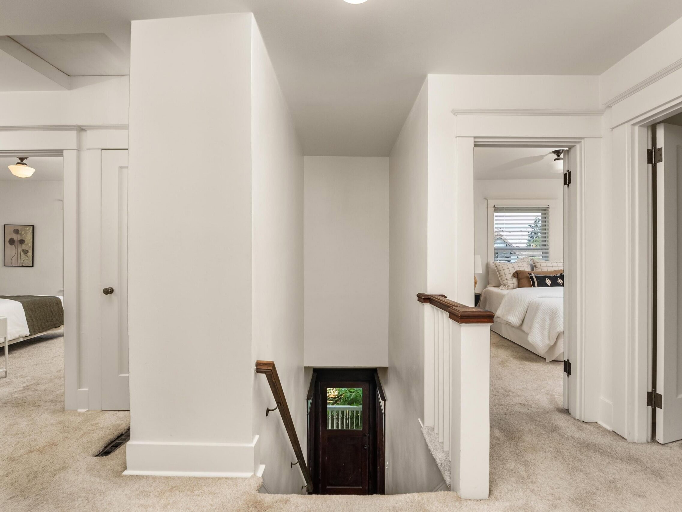 Upstairs hallway with beige carpet, white walls, and doors leading to bedrooms on both sides. A wooden staircase with railing descends to a lower level at the center. Natural light enters from bedroom windows.
