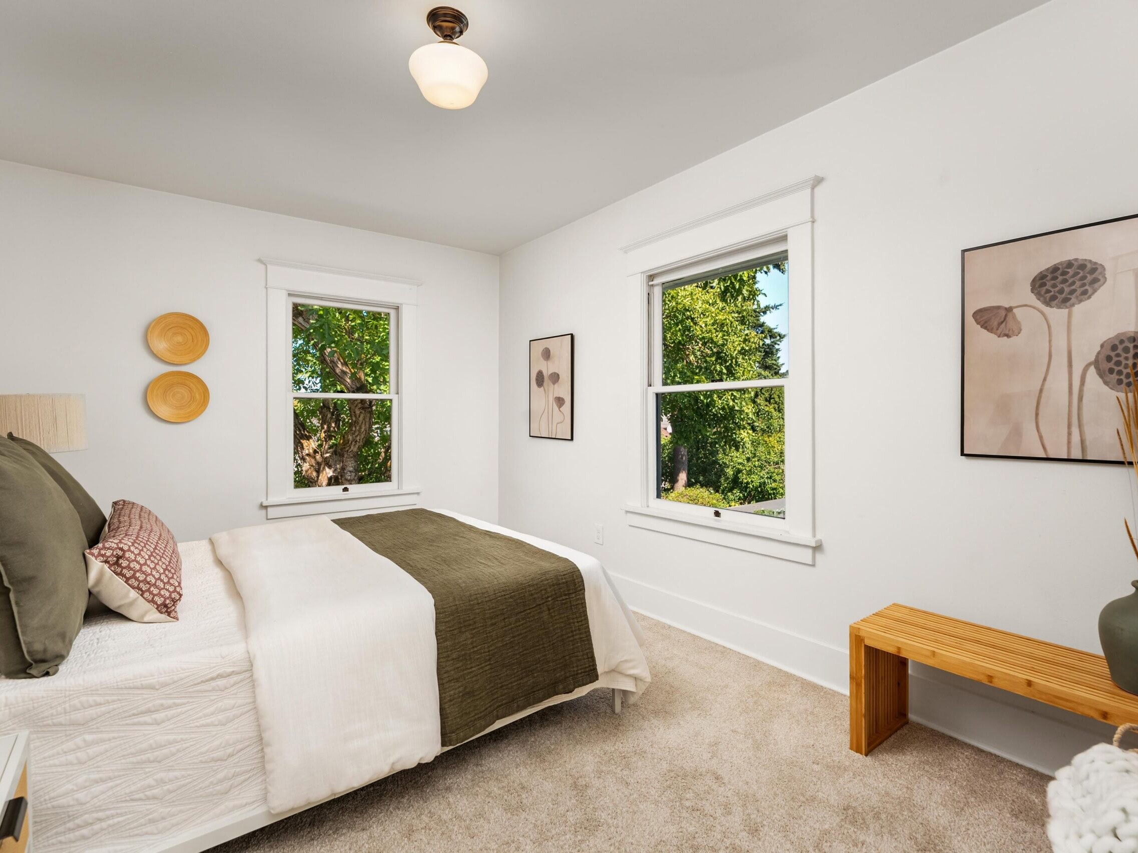 Bright bedroom with a neatly made bed, olive green blanket, two large windows, neutral wall art, a wooden bench, beige carpet, and a vase with decorative sticks on the bench.