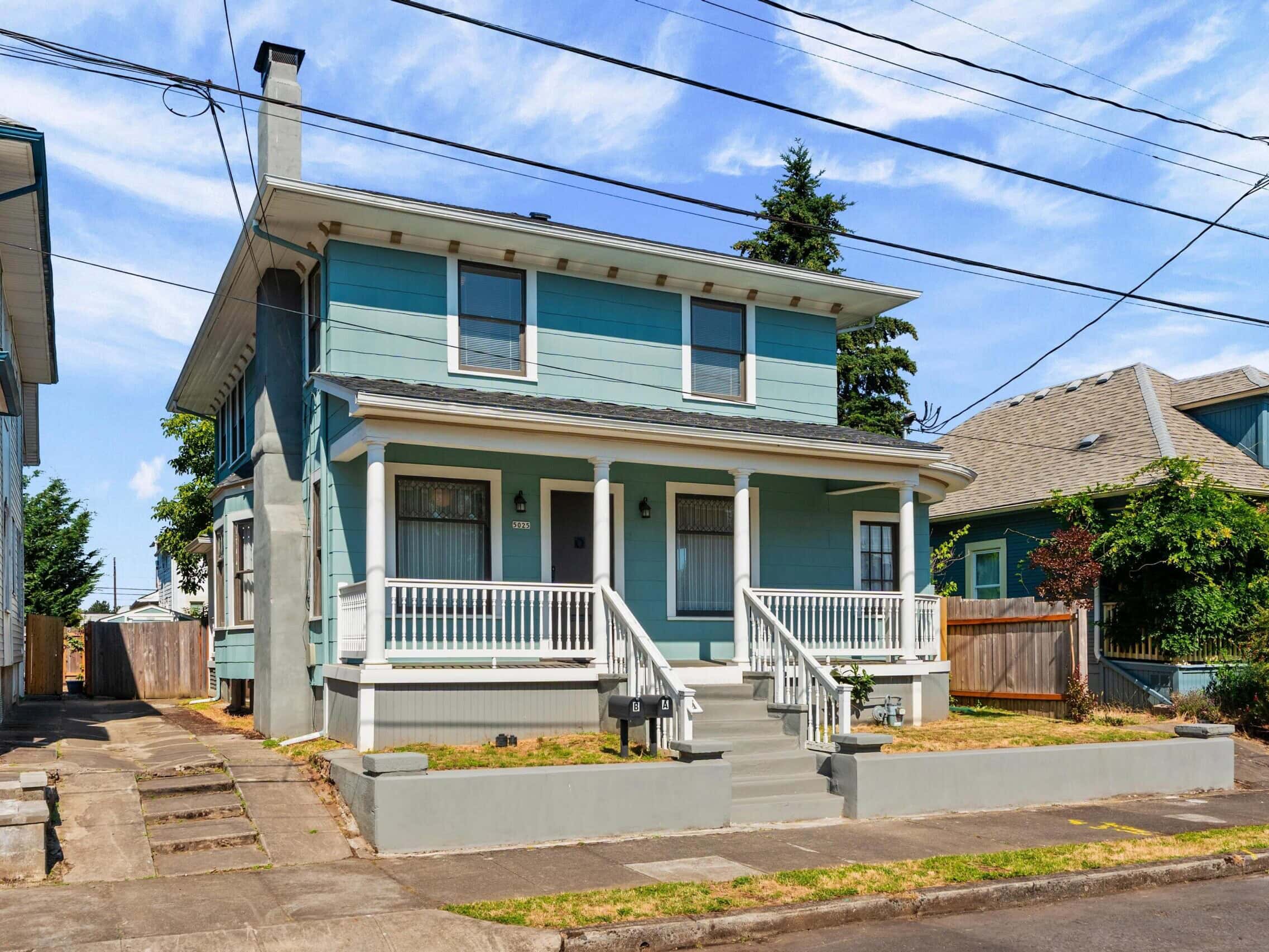 Two-story blue house with white trim, front porch, steps, and railing. Small lawn and concrete walkway. Neighboring homes and tree in background, under a partly cloudy sky.