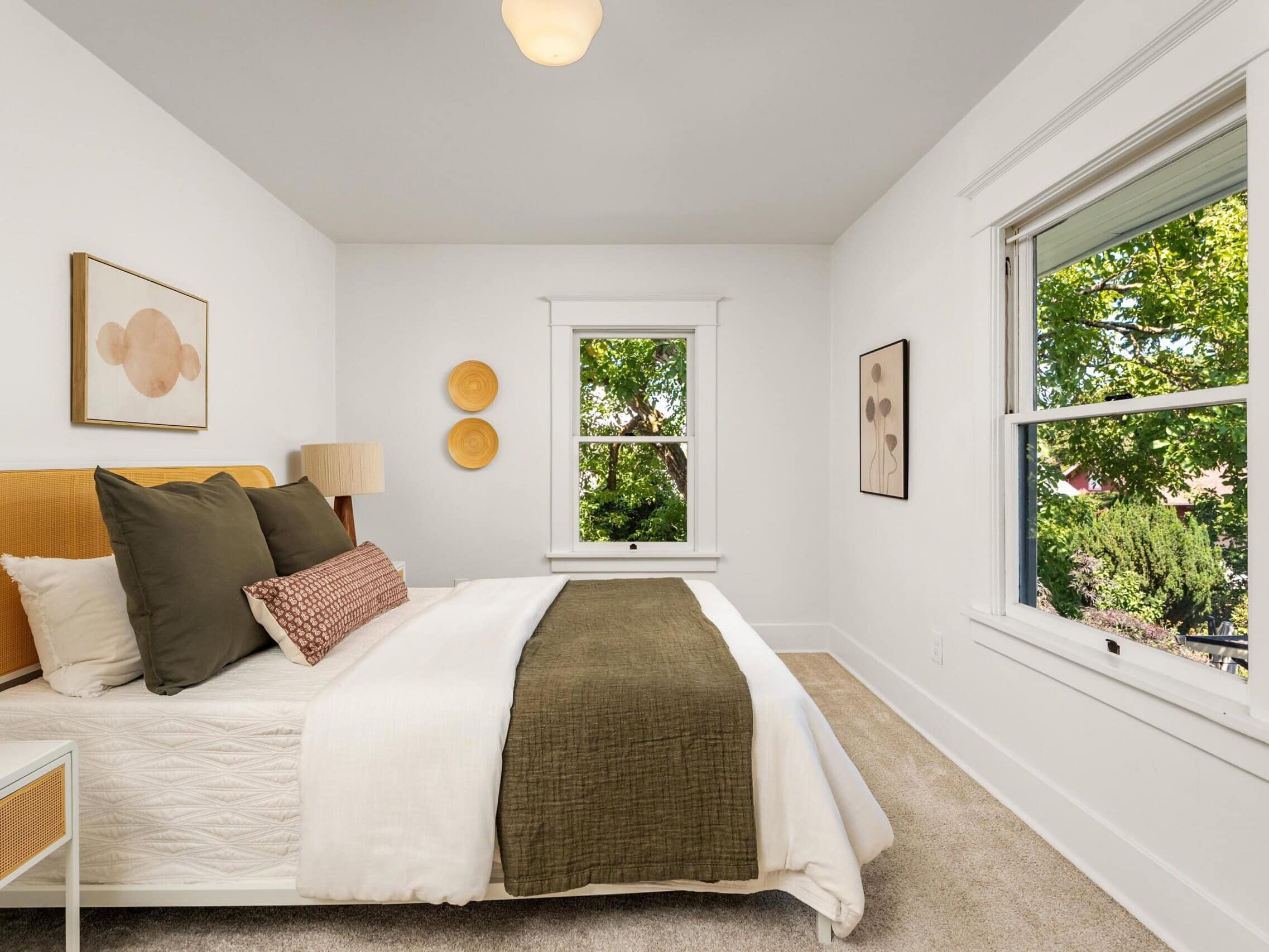 A bright bedroom with a neatly made bed, green and white bedding, two windows, neutral wall art, white walls, and beige carpet, with trees visible outside.
