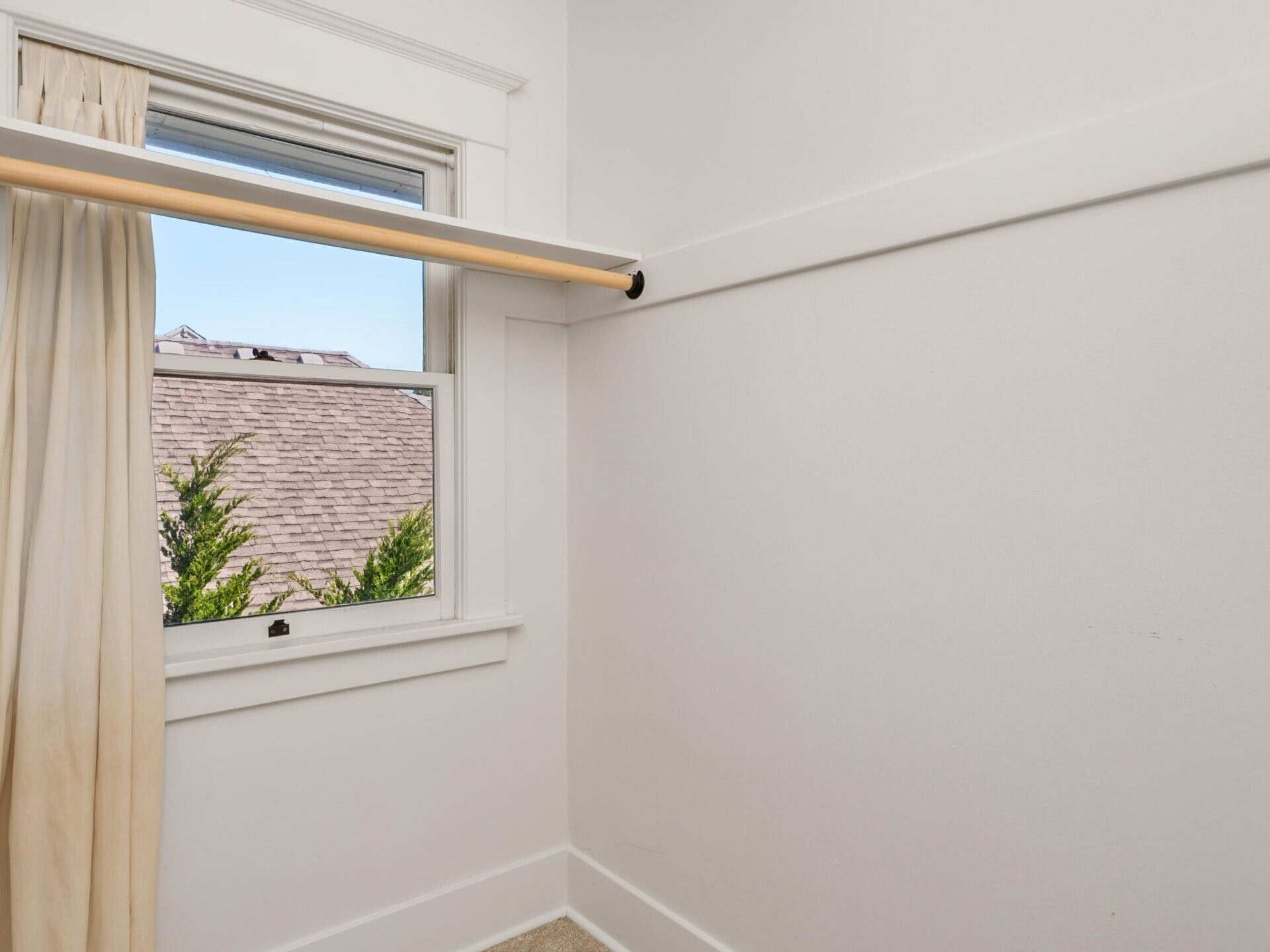 Small empty walk-in closet with beige carpet, white walls, a curtain rod, and a window with a cream curtain. Tree branches and a rooftop are visible outside the window.
