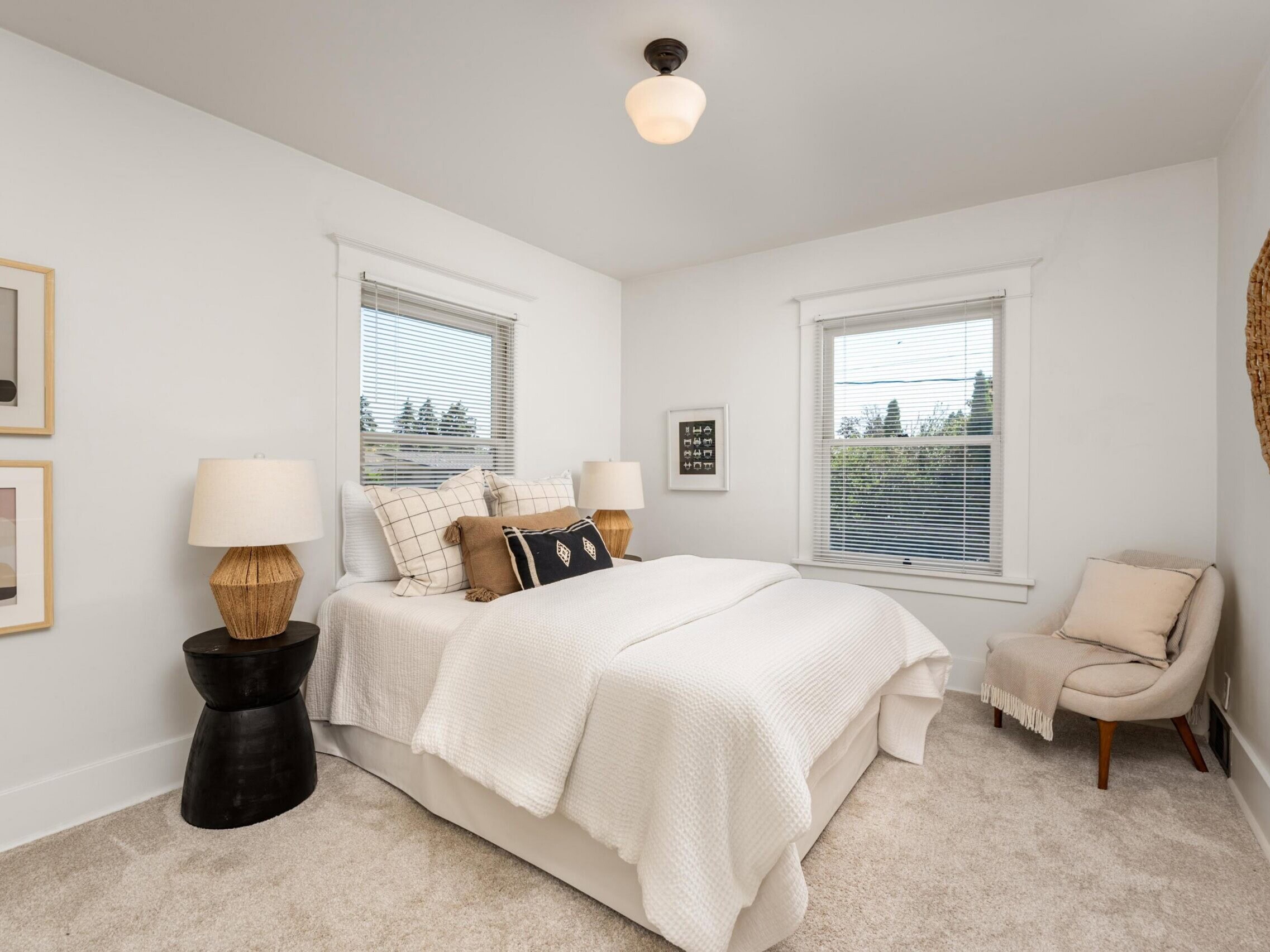 A cozy bedroom with a white bed, neutral pillows, two lamps on side tables, an accent chair, abstract art on the wall, and windows letting in natural light.