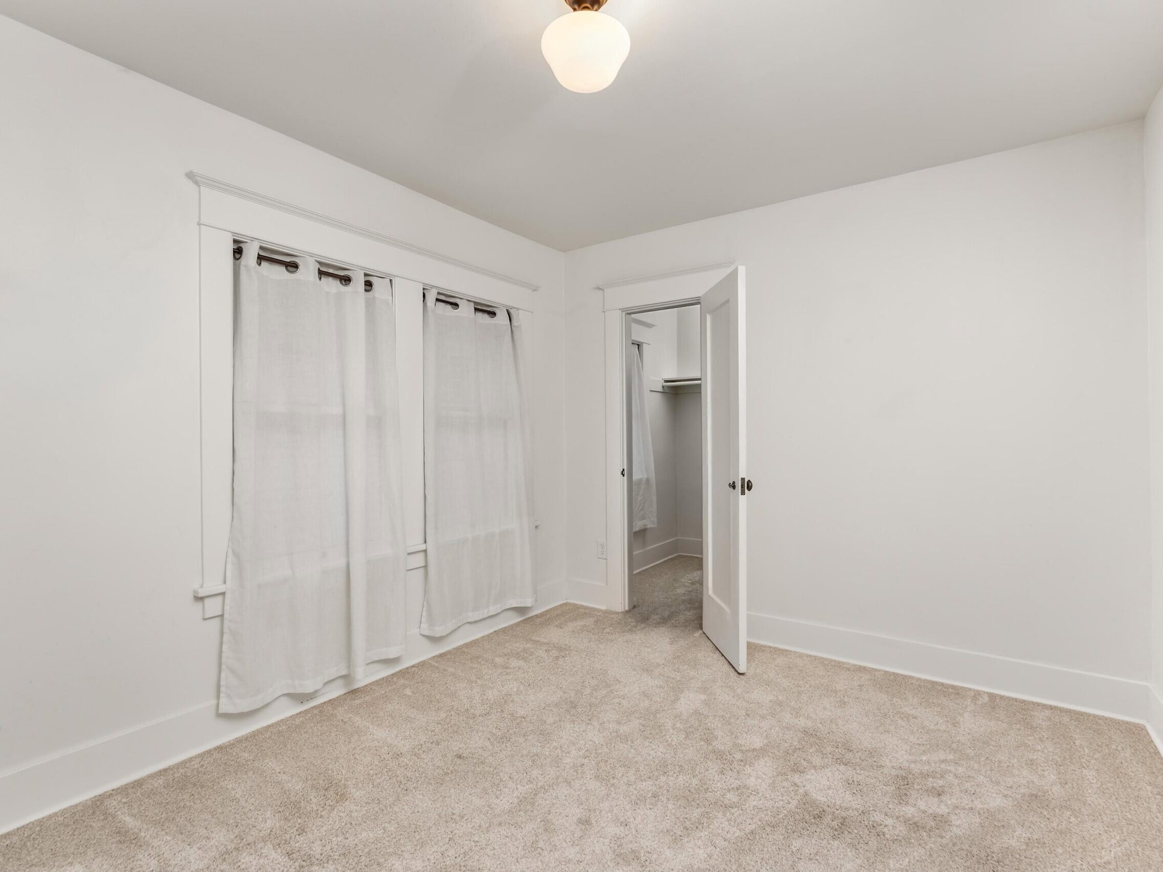 Empty room with beige carpet, white walls, two curtained windows, a ceiling light, and an open door leading to a small walk-in closet.