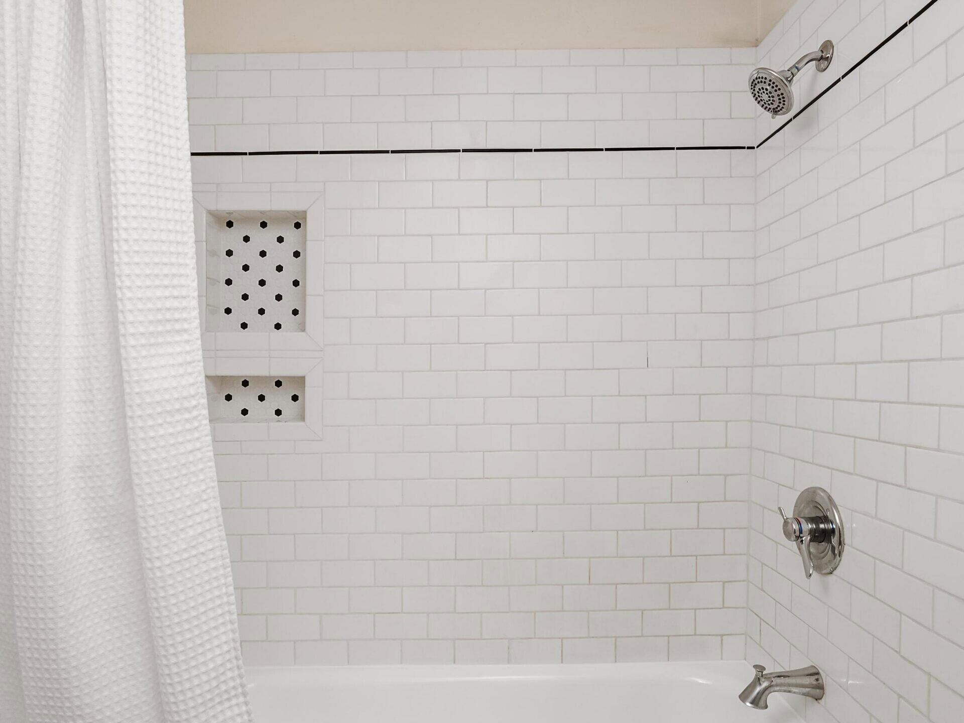 A clean white bathtub with a white tile wall, silver faucet, showerhead, and a white shower curtain. There is a small built-in shelf with toiletries on the left wall.
