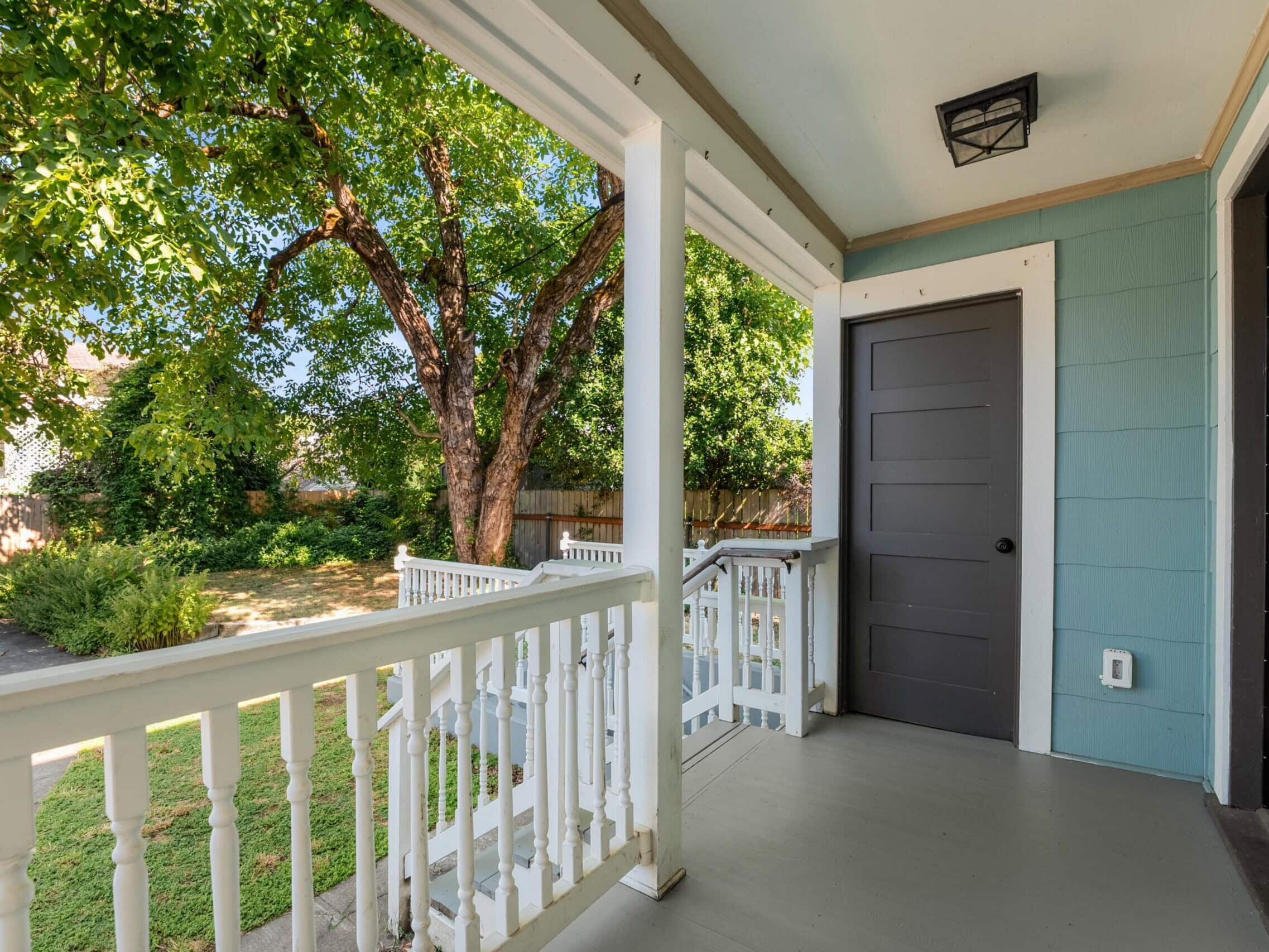 A front porch with white railings, a gray door, light blue siding, and a view of green trees and a walkway leading to the street.
