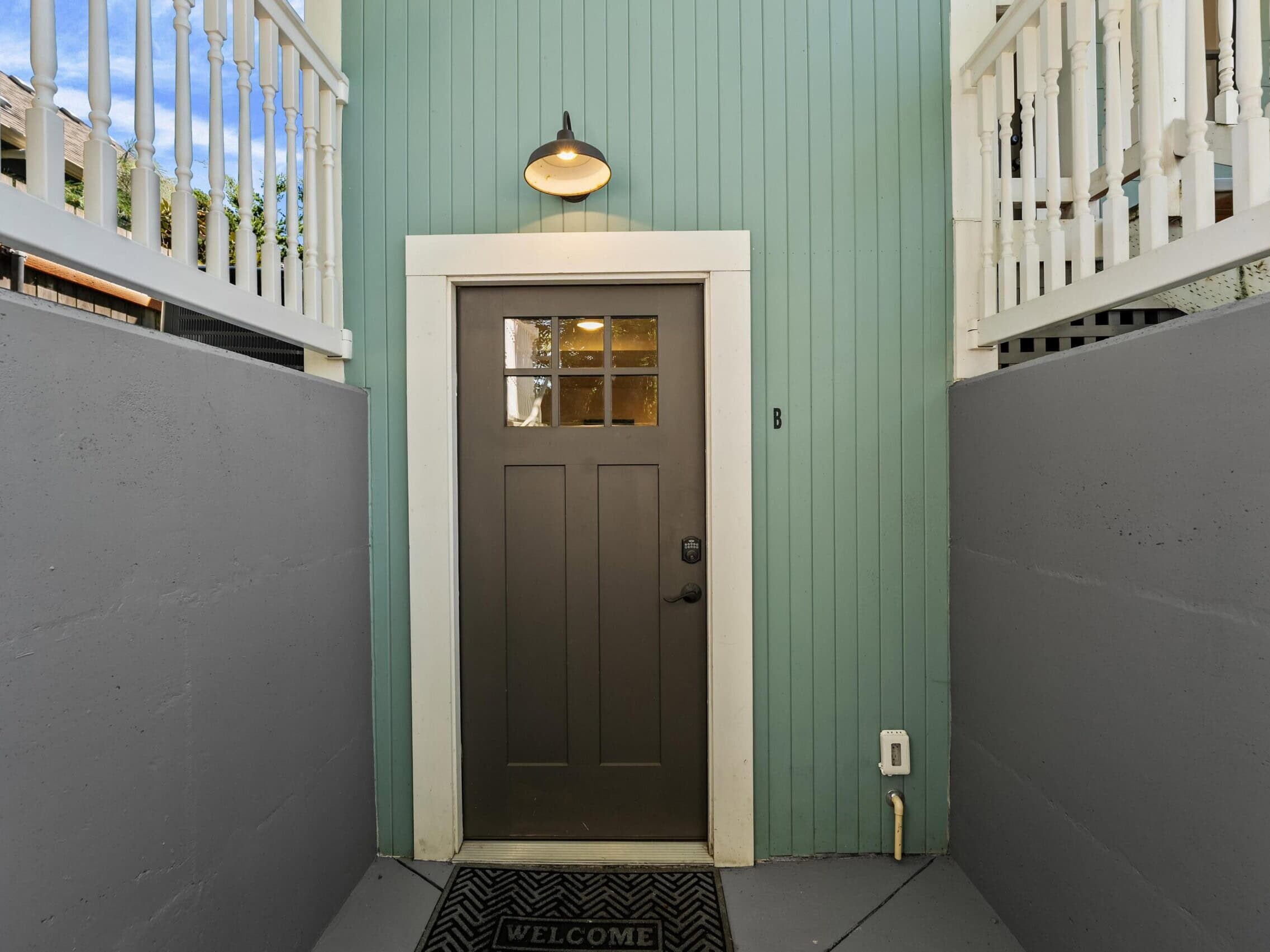 A gray door with a window and a B sign is set in a green wall, flanked by white railings and concrete walls. A light fixture is above the door and a WELCOME mat is on the ground.