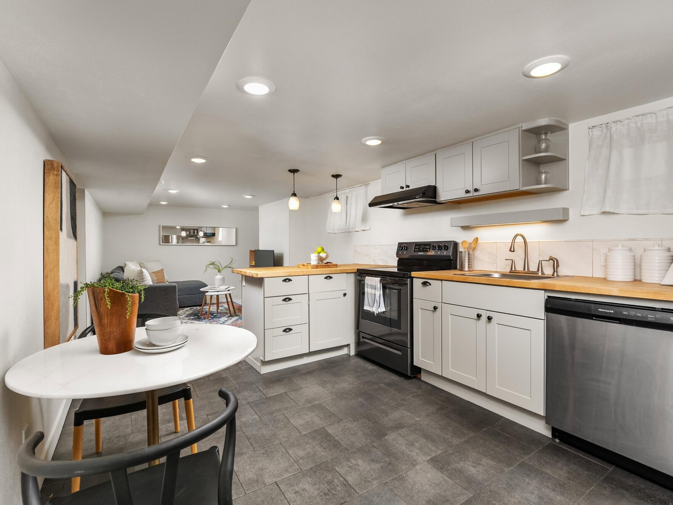 Modern kitchen with white cabinets, wood countertops, stainless steel appliances, a small round dining table with two chairs, and cozy living area in the background under recessed lighting.