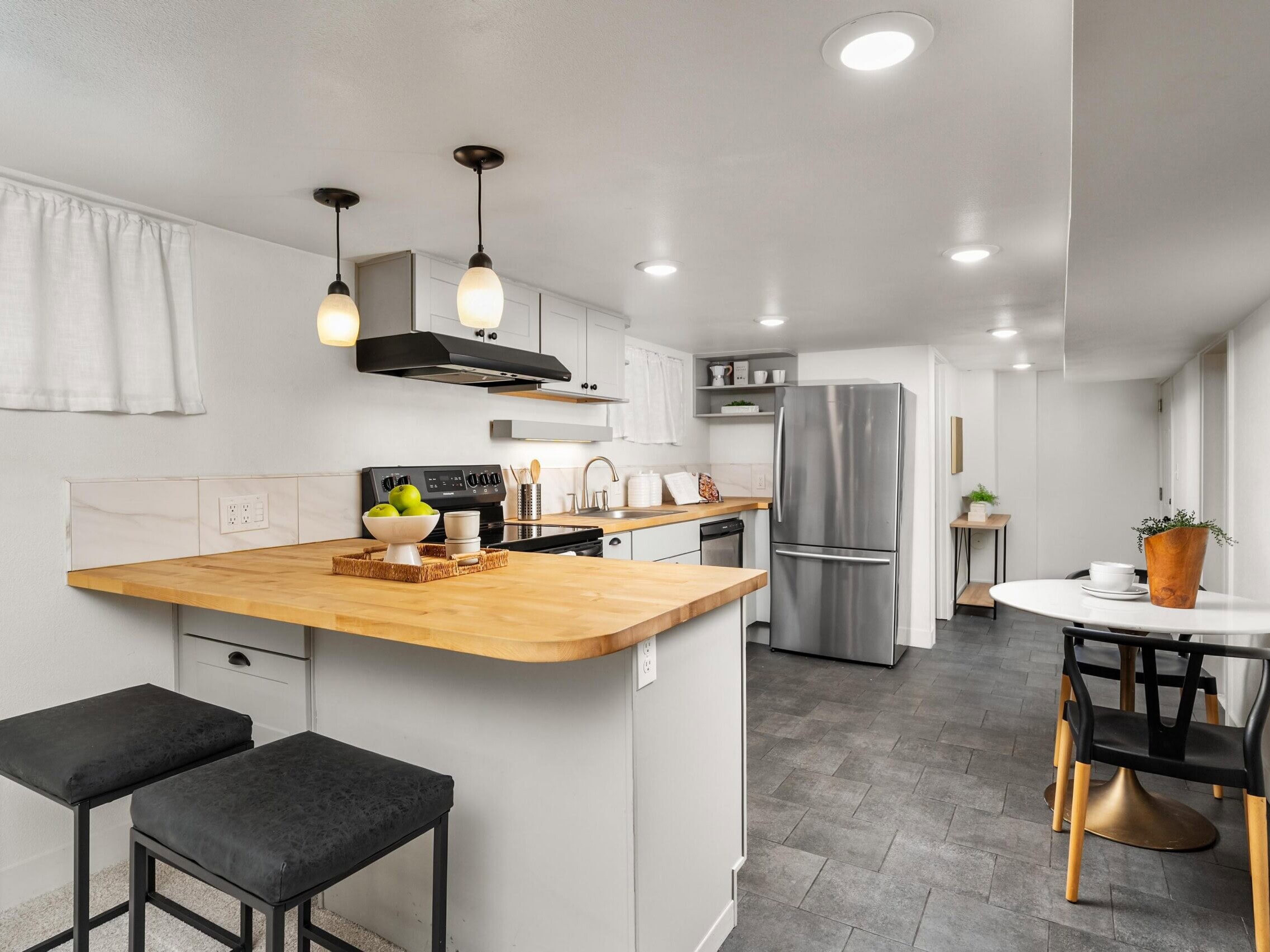 Modern kitchen with light wood countertops, white cabinets, stainless steel appliances, black stools, pendant lights, and a small round dining table with two chairs and a potted plant by the window.