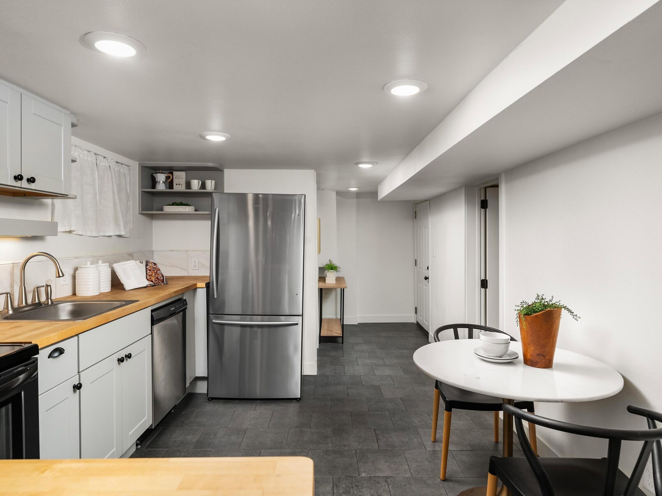 Modern kitchen with stainless steel appliances, white cabinets, wood countertops, a small round dining table with two chairs, and a potted plant. The room has gray tile flooring and bright overhead lighting.