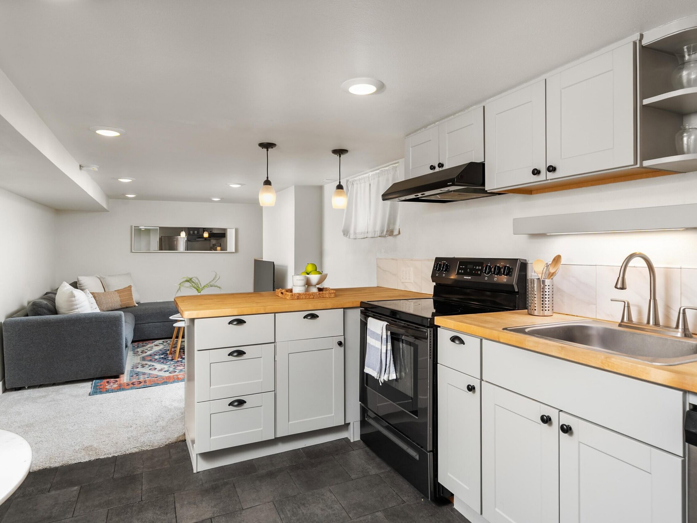 Modern kitchen with light gray cabinets, wood countertops, and black appliances, open to a cozy living area with a gray sofa, white walls, and recessed lighting.
