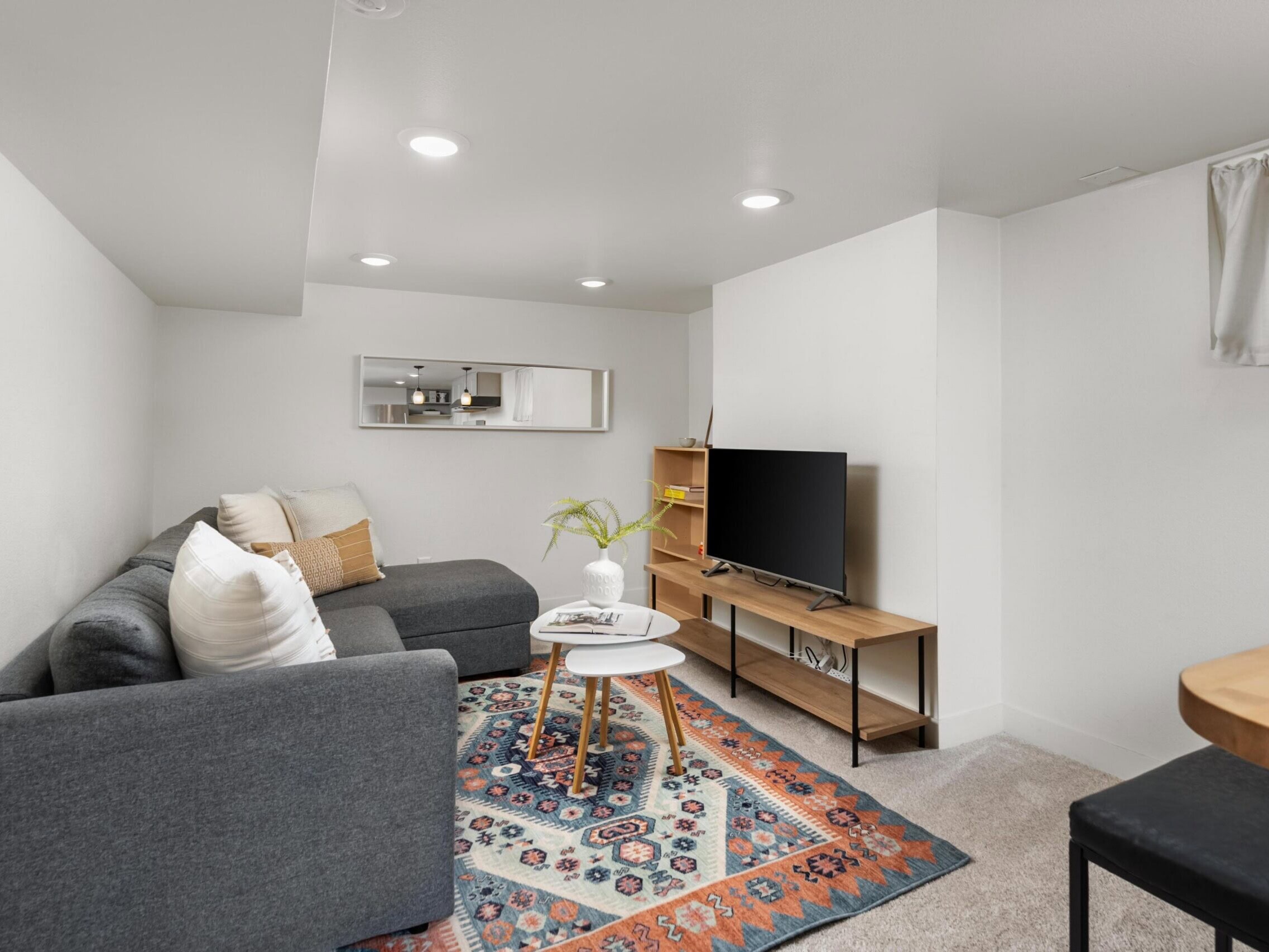 Cozy living room with a gray sectional sofa, two round tables, TV on a wooden stand, colorful rug, shelves, and a mirror on the white wall. Bright lighting and minimal decor create a modern, inviting space.
