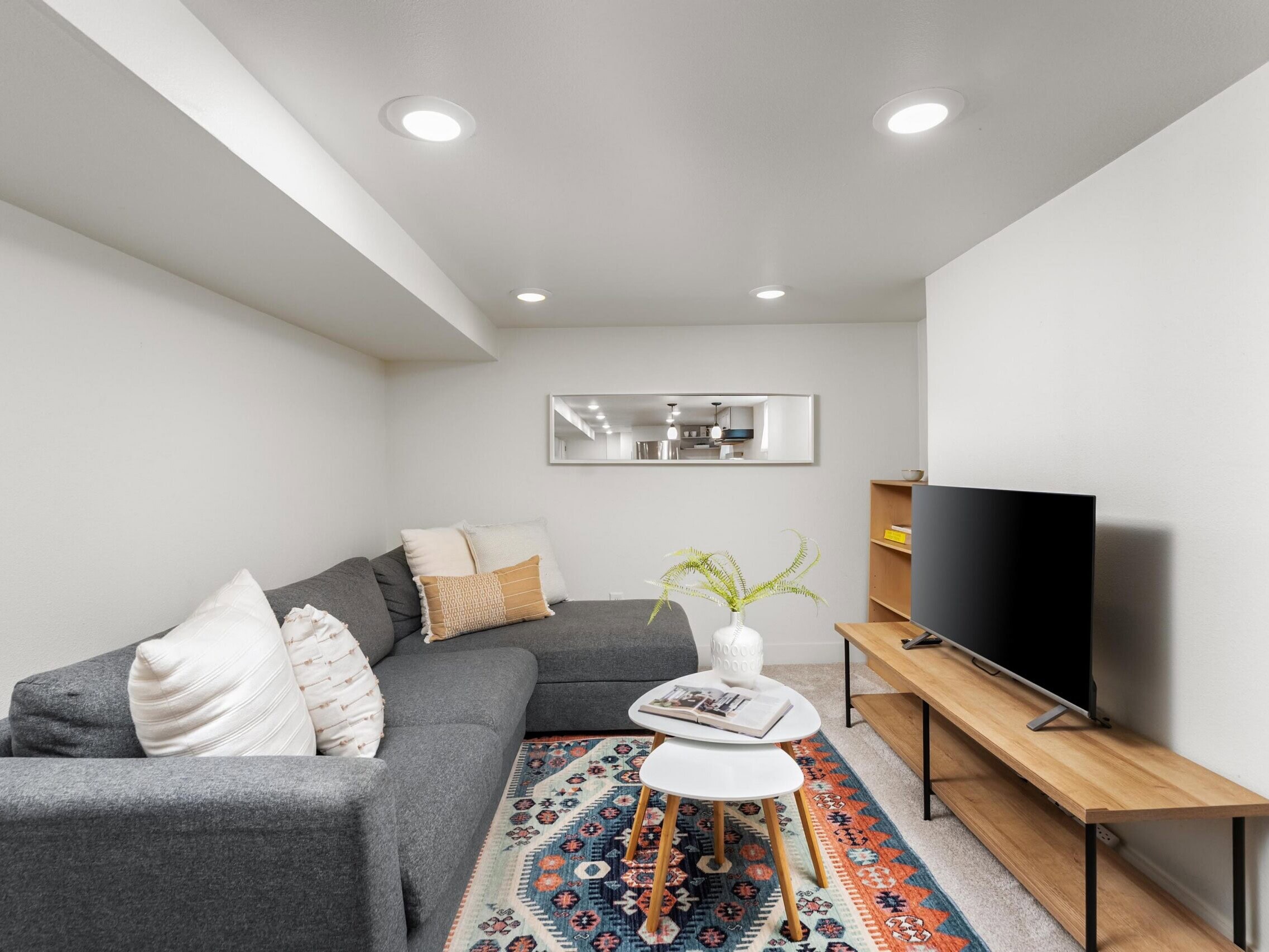 A modern living room with a gray sectional sofa, colorful rug, wooden TV stand, flat-screen TV, small tables, and a wall mirror reflecting shelves and decor. White walls and ceiling with recessed lighting.