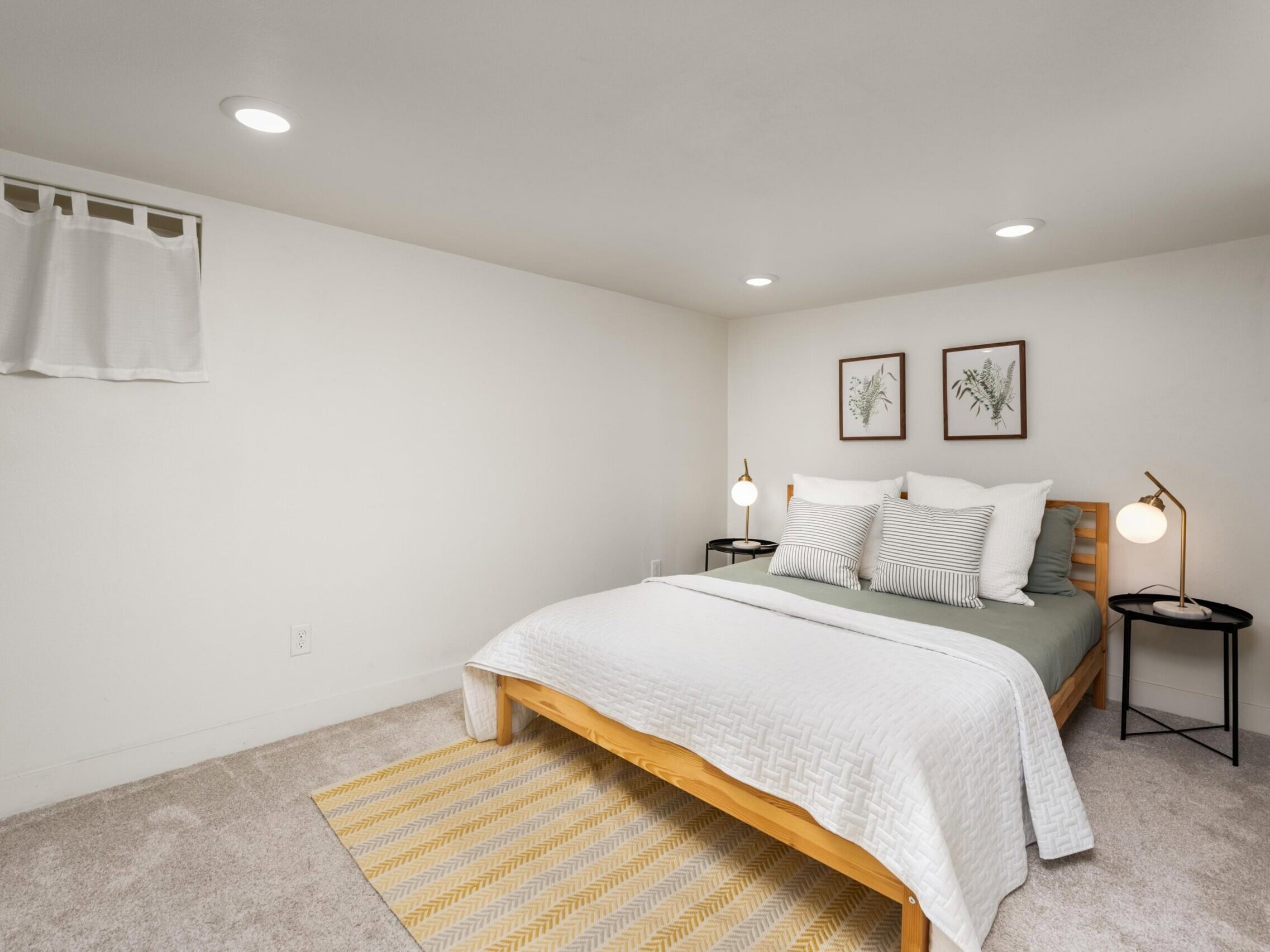 Minimalist bedroom with a wooden bed, white bedding, green pillows, two side tables with lamps, two botanical prints on the wall, and a small window with a white curtain. Light beige carpet and a yellow rug.