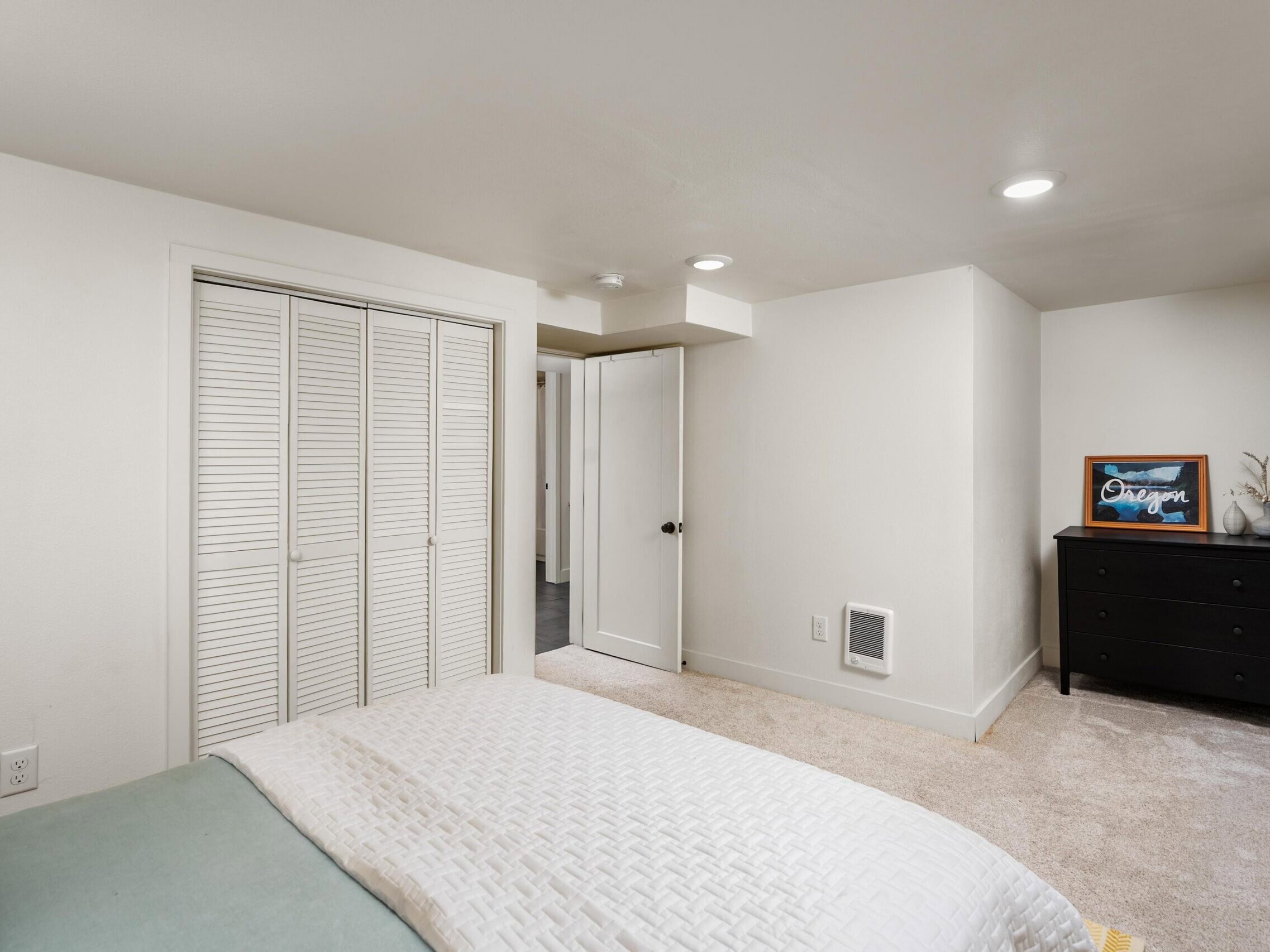 A bedroom with a neatly made bed, white closet doors, beige carpet, a black dresser with a framed picture, and a slightly open door leading to another room.