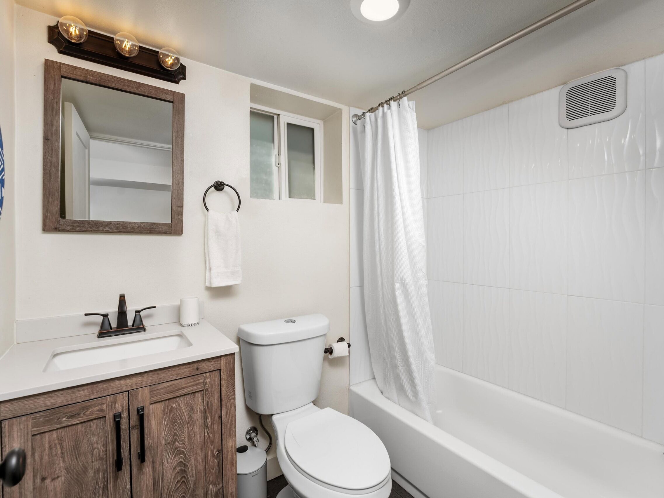Modern bathroom with a wooden vanity, rectangular mirror, toilet, white shower curtain, and white tile surround. Three light bulbs are above the mirror. A towel and toilet paper are on holders near the sink and toilet.