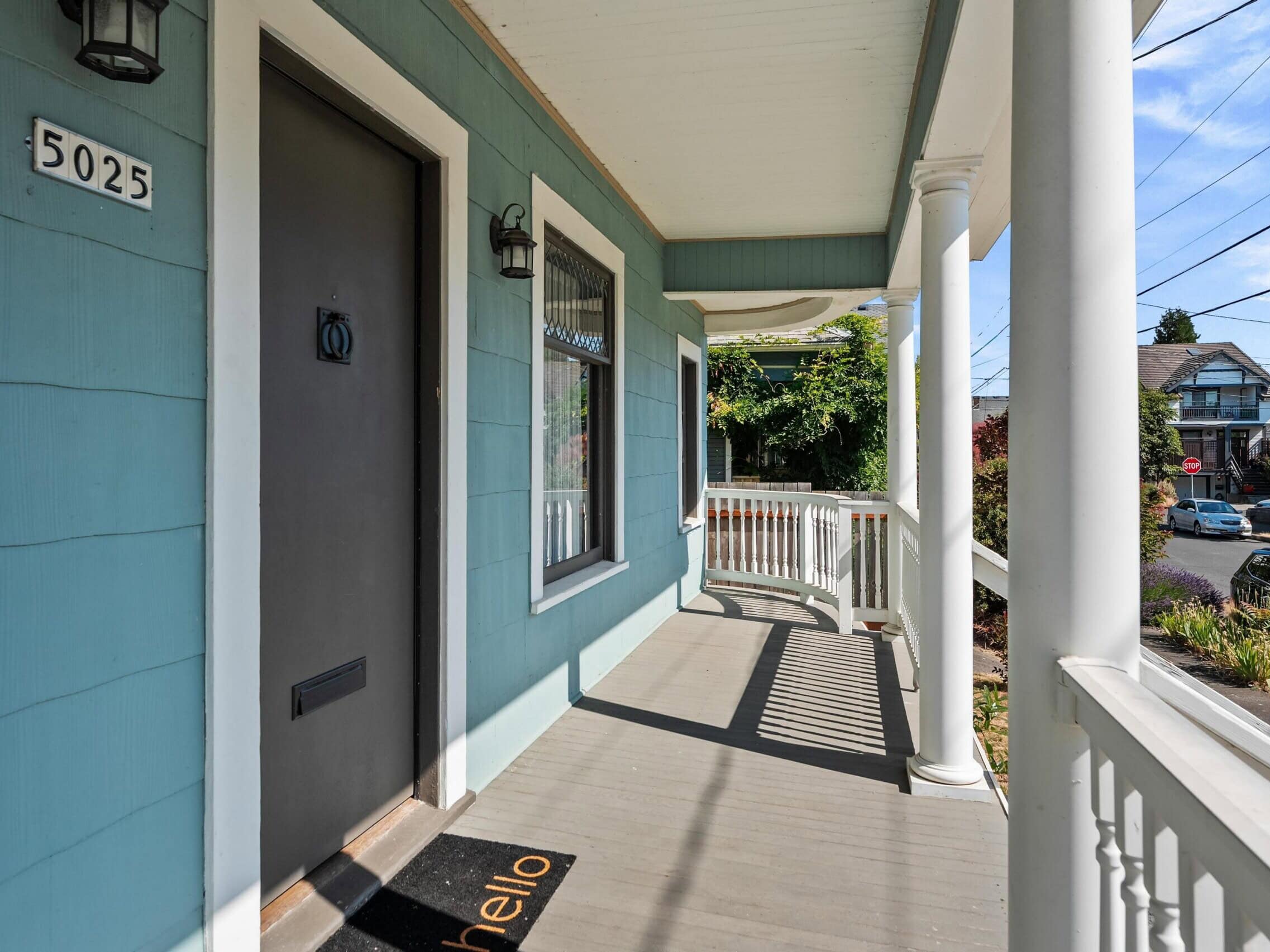 A blue house porch with white railing and columns, a black door with the number 5025, a hello doormat, and a sunny street view with neighboring houses and greenery.
