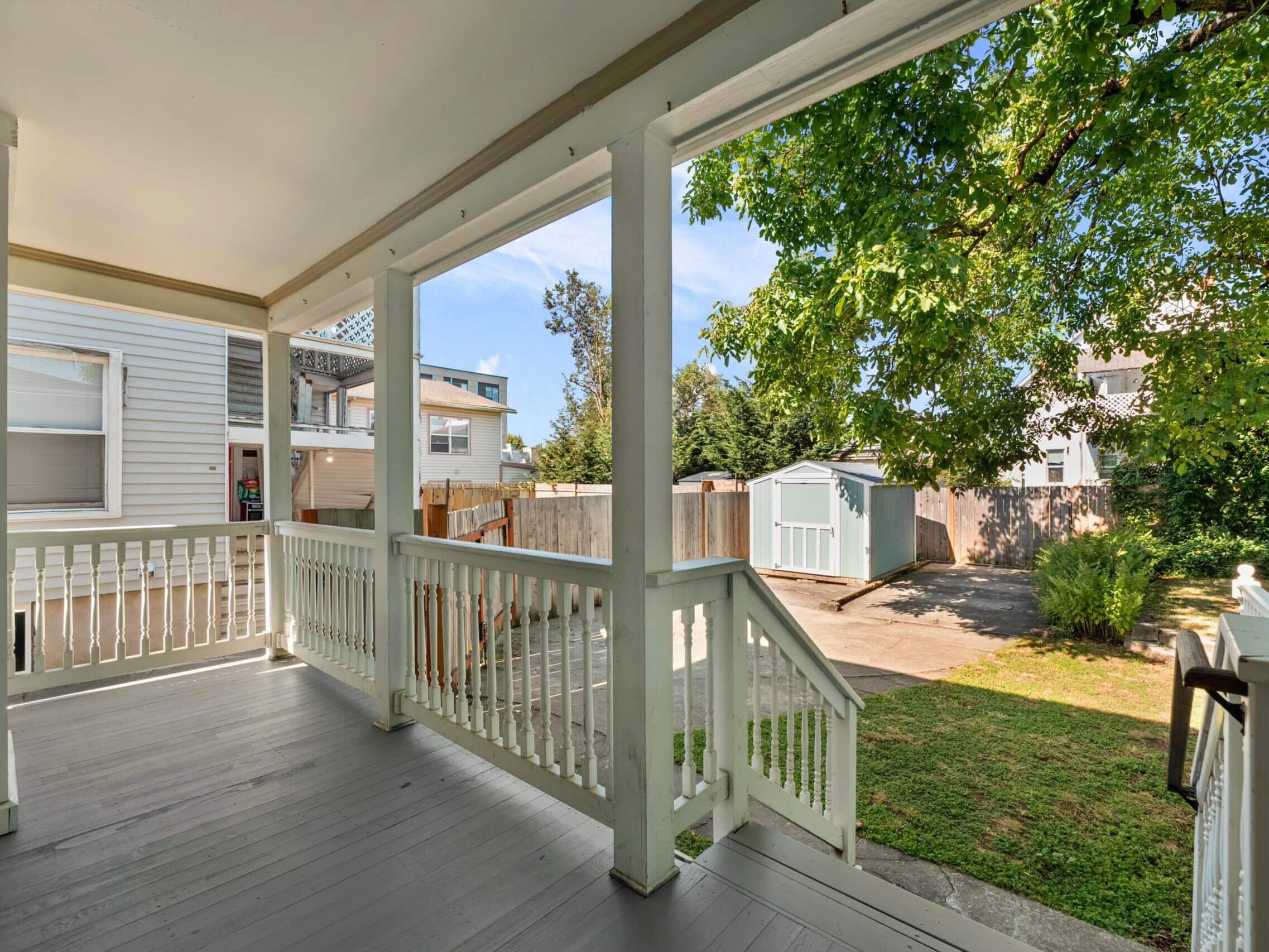 A covered porch with white railings overlooks a backyard with green grass, a tree, and two sheds. Neighboring houses and a clear sky are visible in the background.