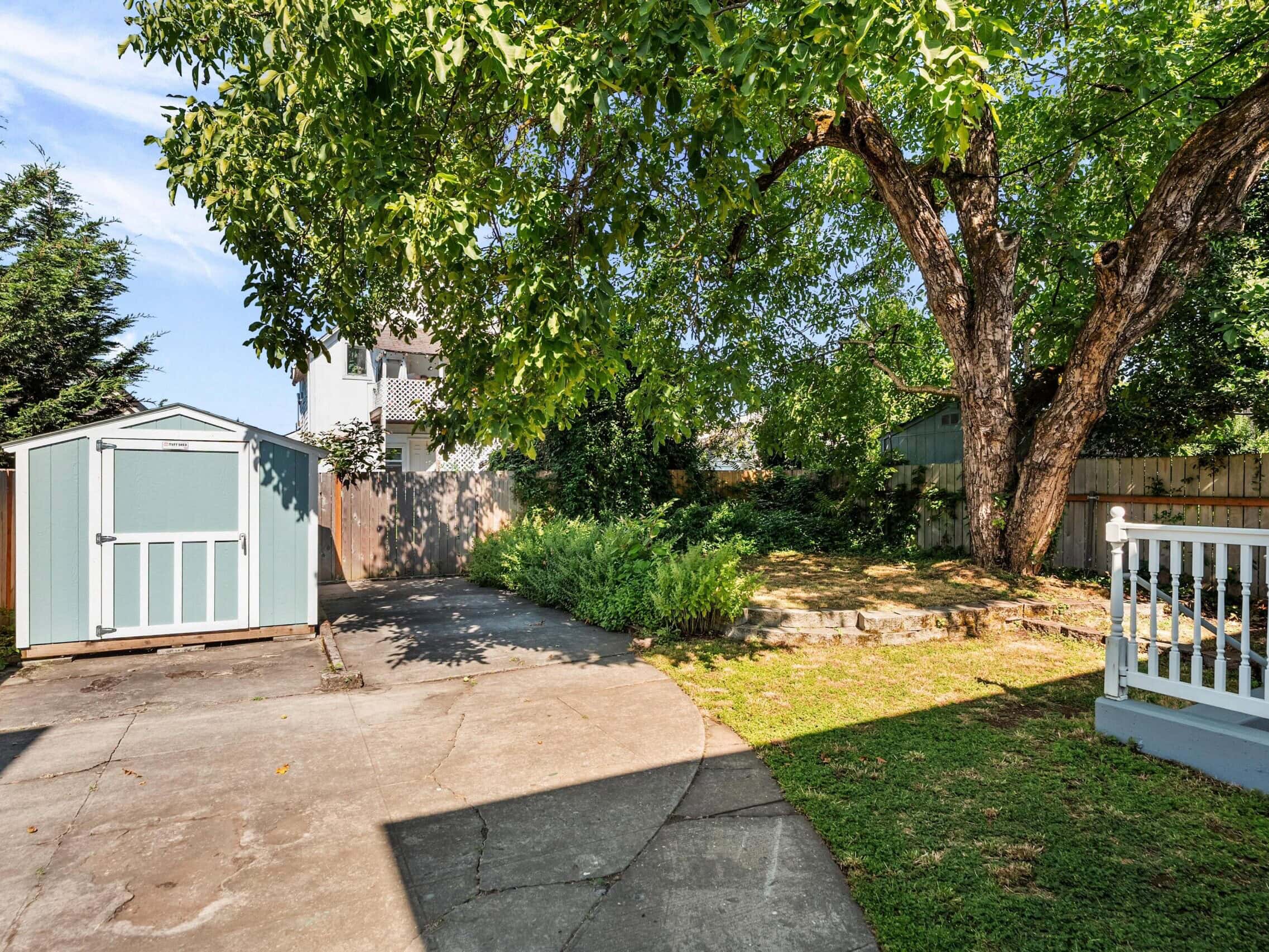 A backyard with a shed on the left, a large tree providing shade, a small garden, and a white-railed porch on the right. The area is partly grass and partly concrete.