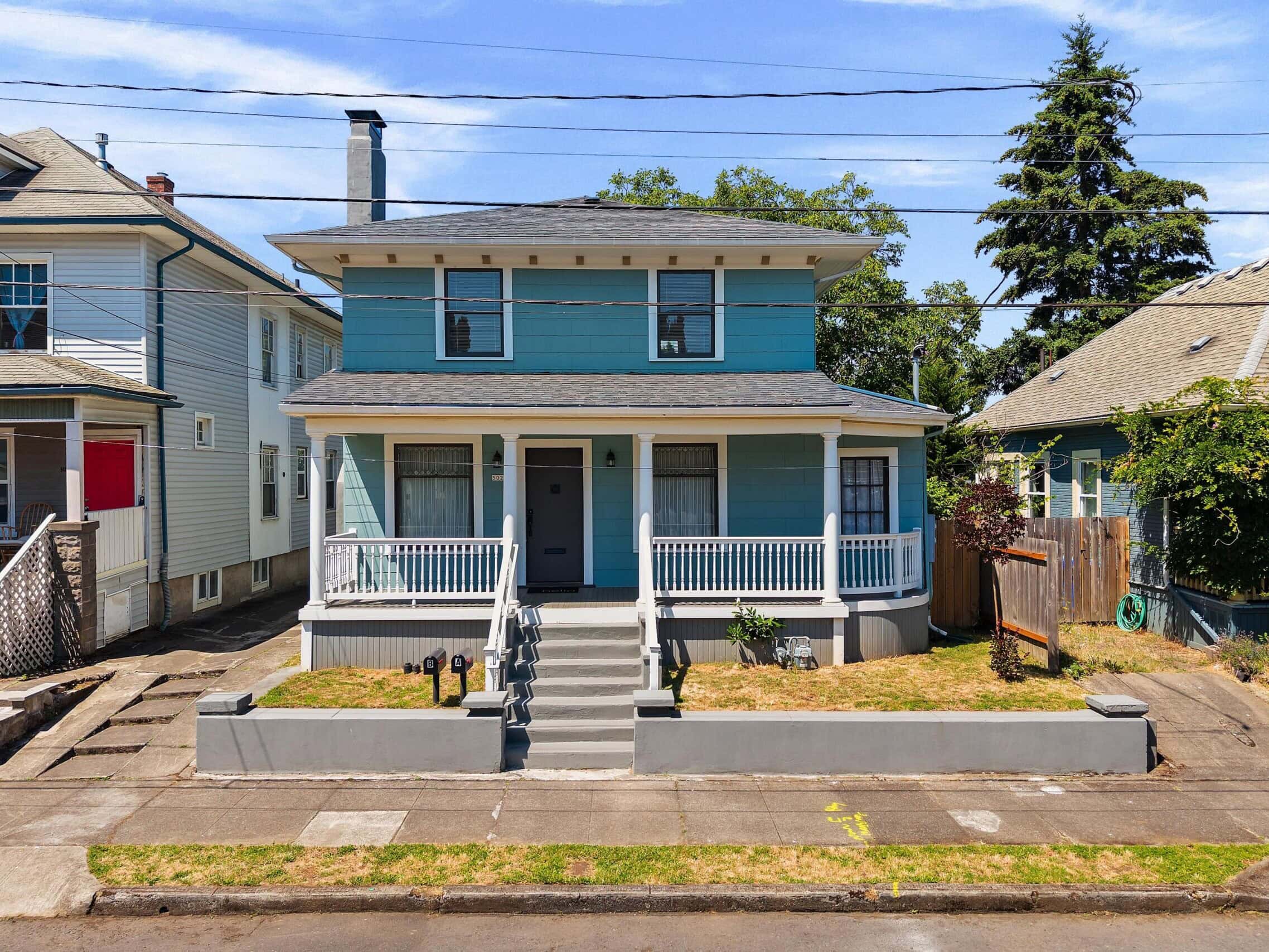 A two-story blue house with white trim, a covered front porch, and steps leading to the entrance. The house sits between two neighboring homes and has a small front yard enclosed by a low gray wall.