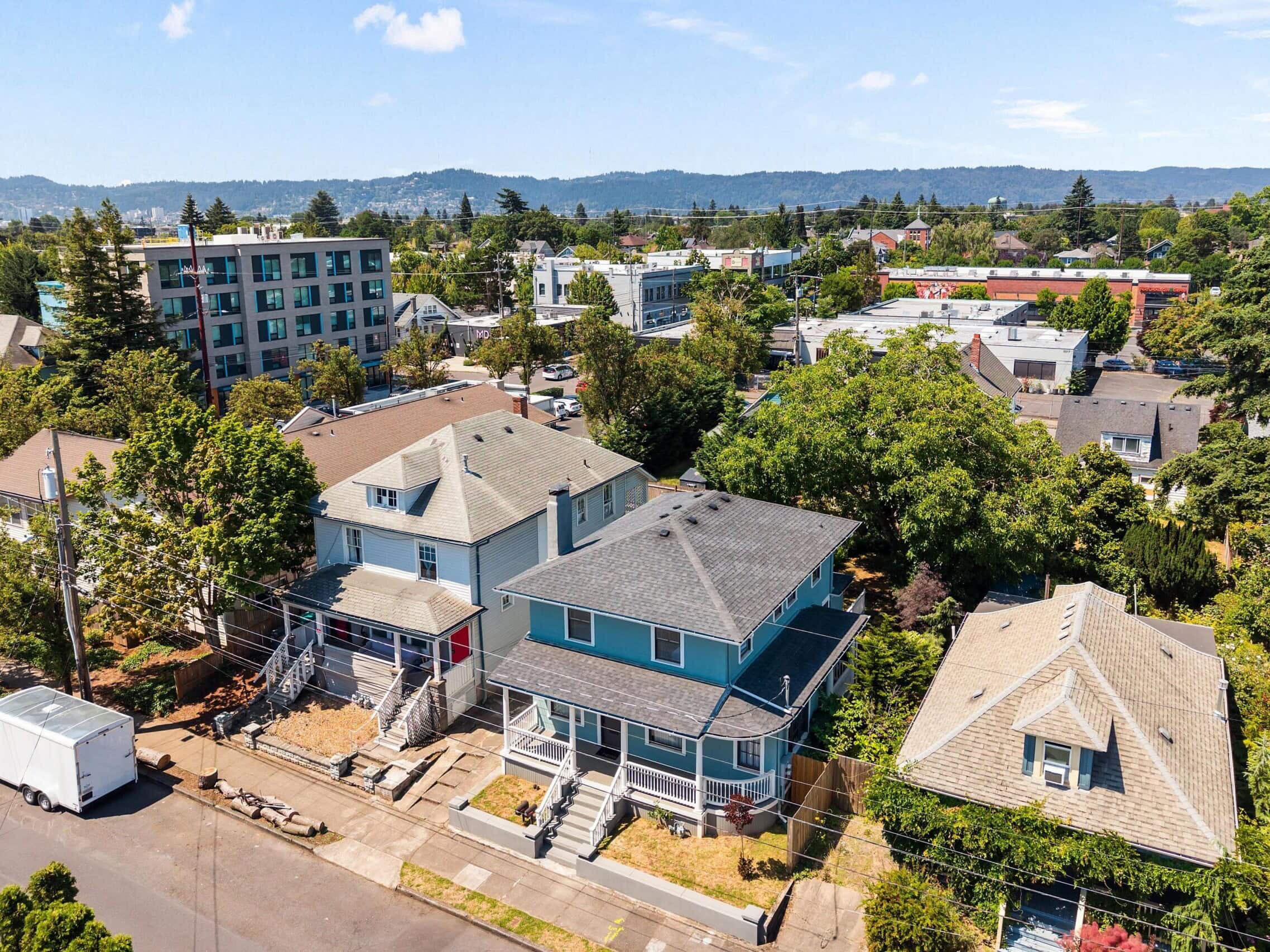 Aerial view of suburban neighborhood with several houses, trees, and apartment buildings under a blue sky with clouds and distant hills in the background.