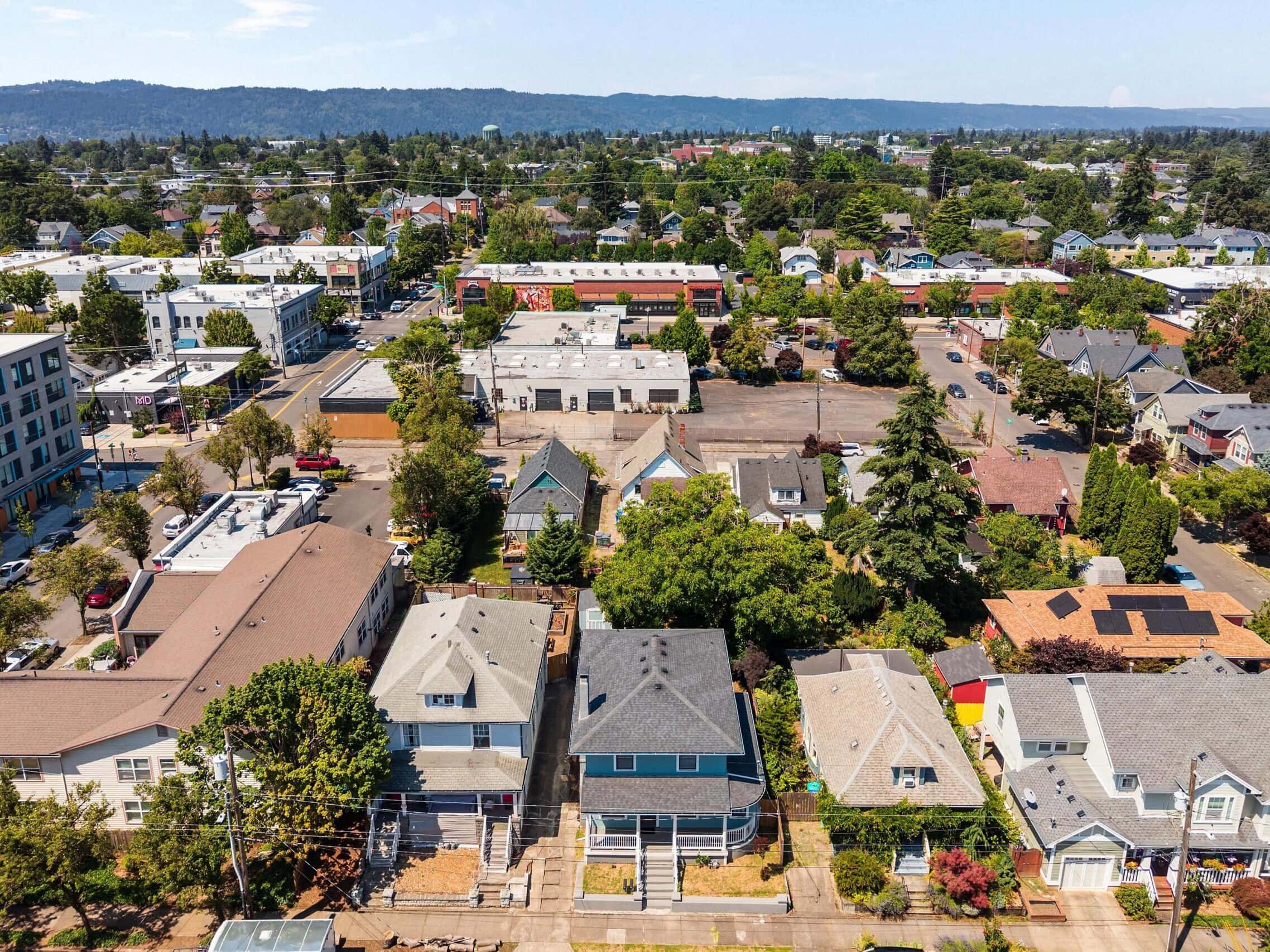 Aerial view of a suburban neighborhood with houses, trees, and streets, and distant hills under a blue sky.