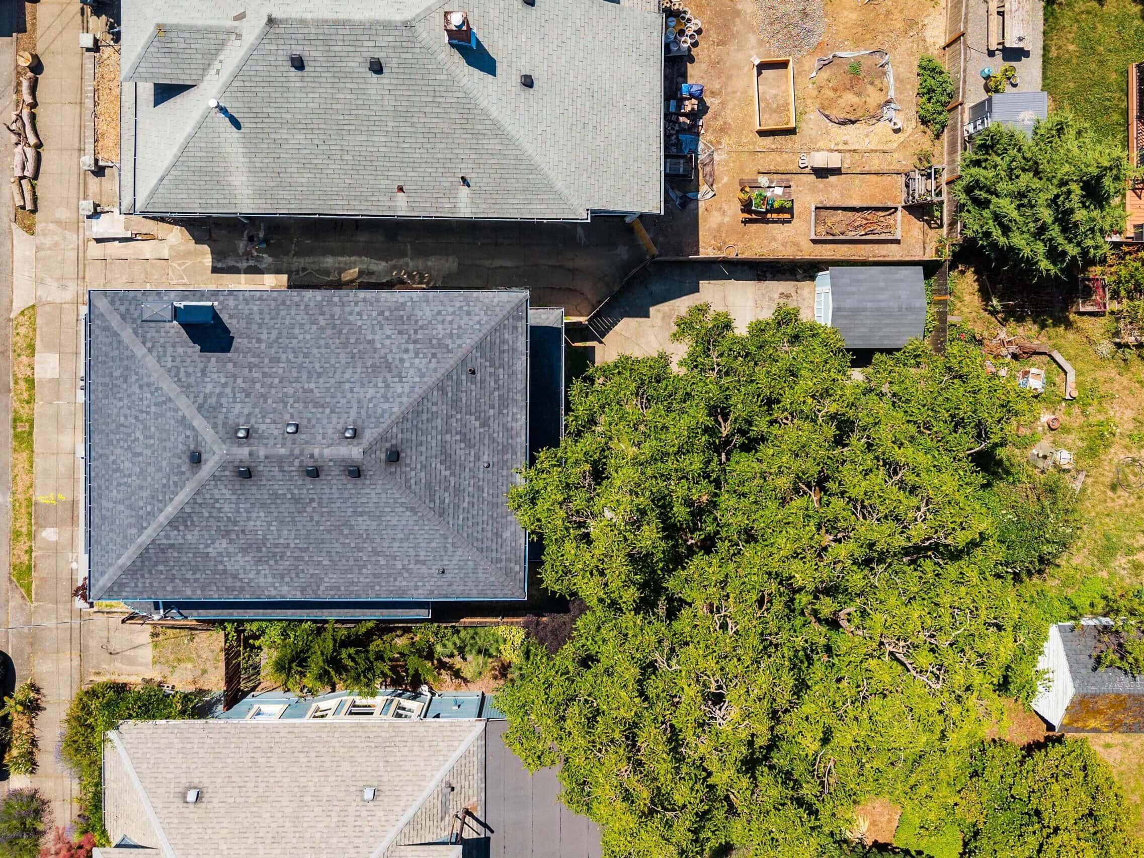 Aerial view of suburban houses, a tree, driveways, a parked car, and backyards with garden beds and patios on a sunny day.