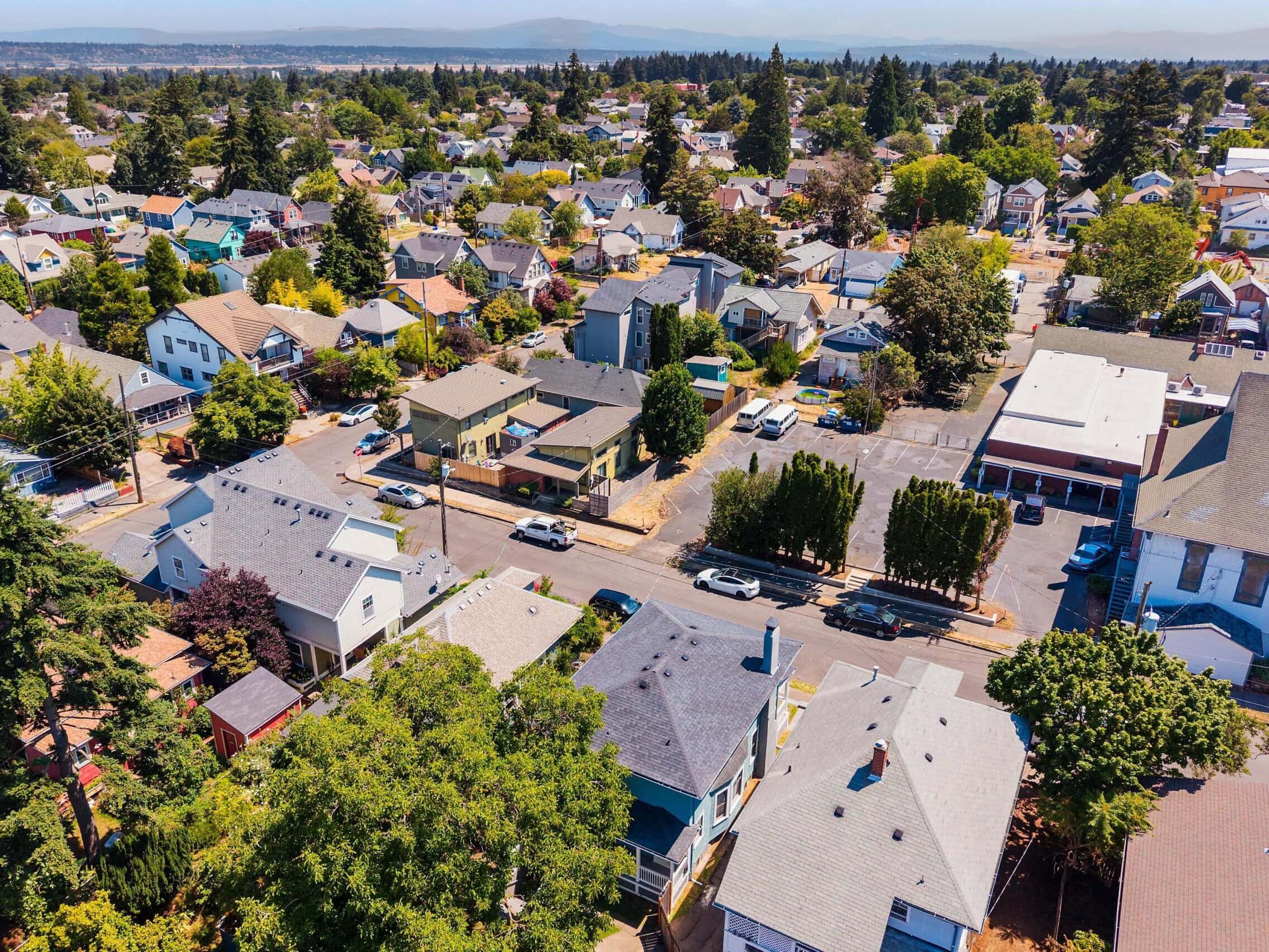 Aerial view of a suburban neighborhood with tree-lined streets, houses, parked cars, and greenery on a sunny day. Distant hills are visible in the background.