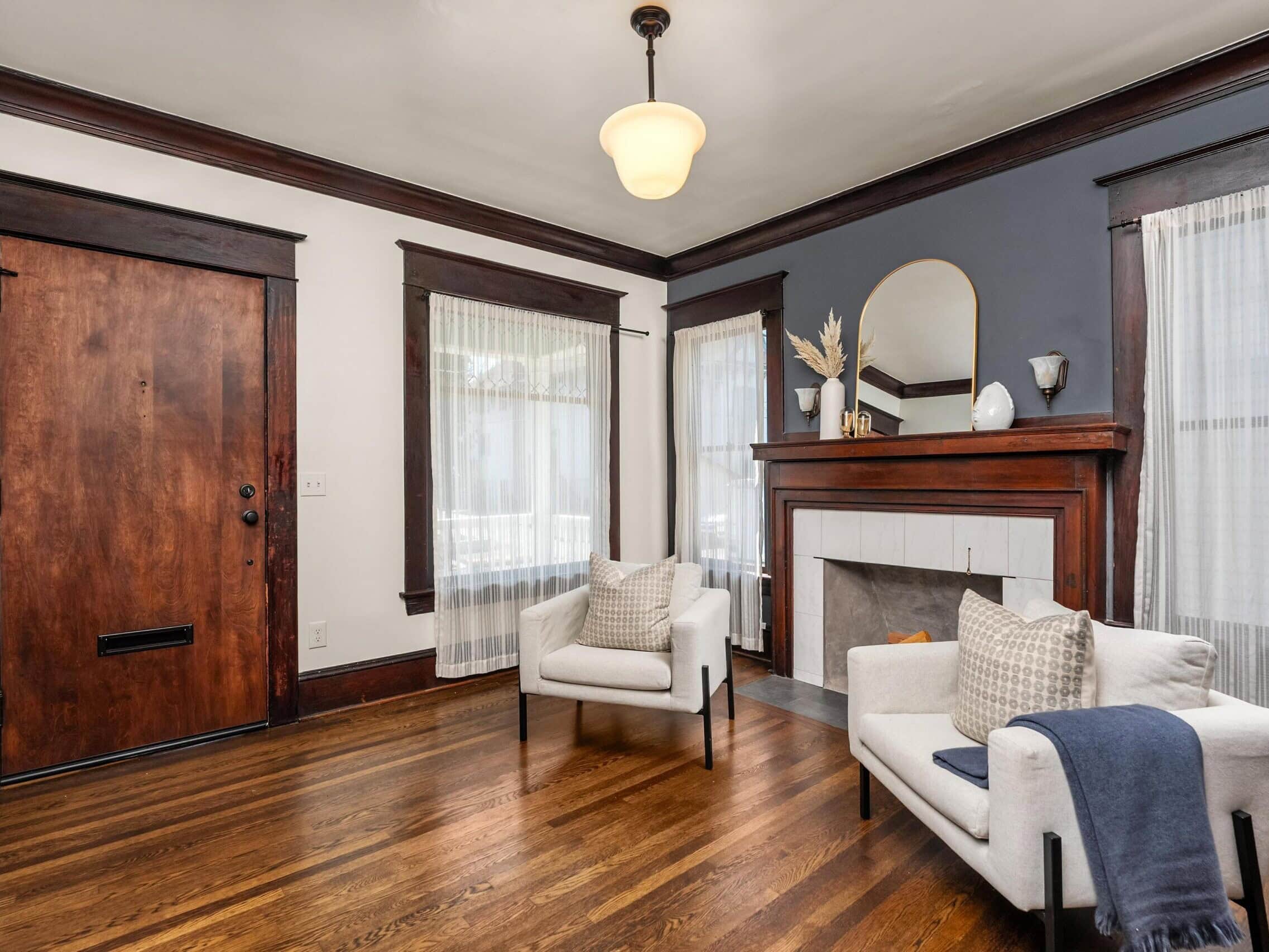 Cozy living room with hardwood floors, two white armchairs, a wooden door, large windows with sheer curtains, and a dark-accented fireplace decorated with vases and dried plants.