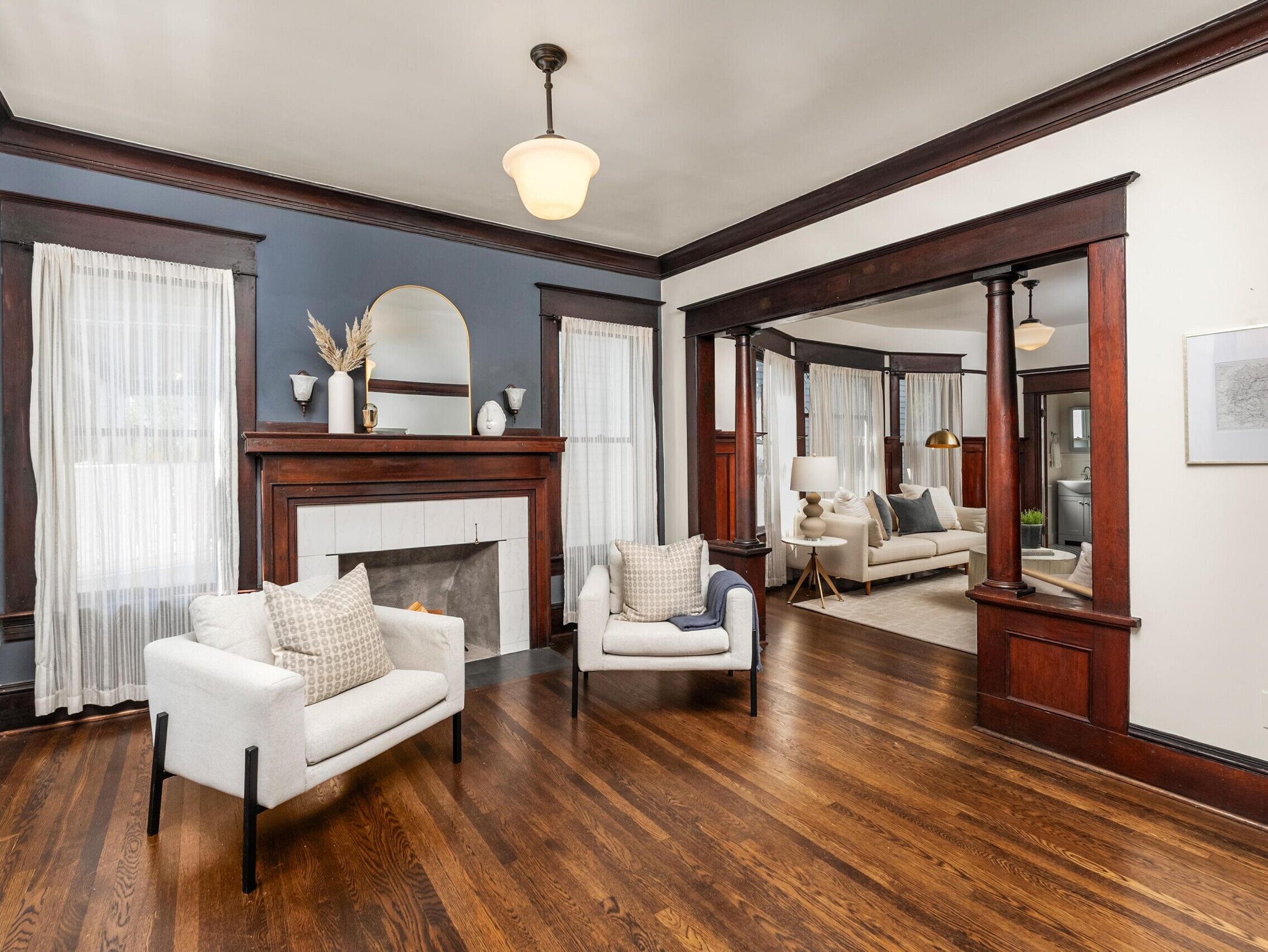 Living room with dark wood trim, hardwood floors, two white armchairs, a fireplace with a mirror above it, and sheer curtains on tall windows. An adjoining room with a sofa is visible in the background.