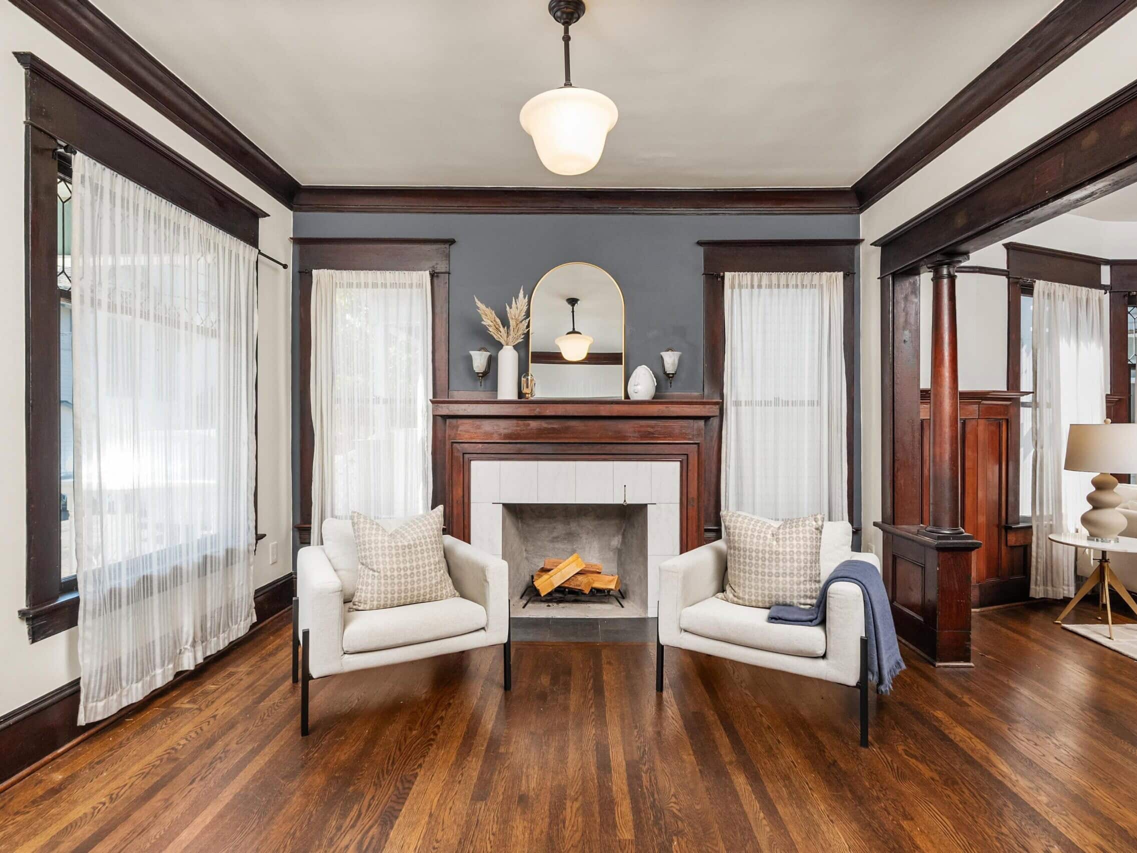 Elegant living room with wooden floors, a dark wood fireplace, two light armchairs, and sheer curtains. A round mirror and decor sit on the mantle under a modern light fixture.