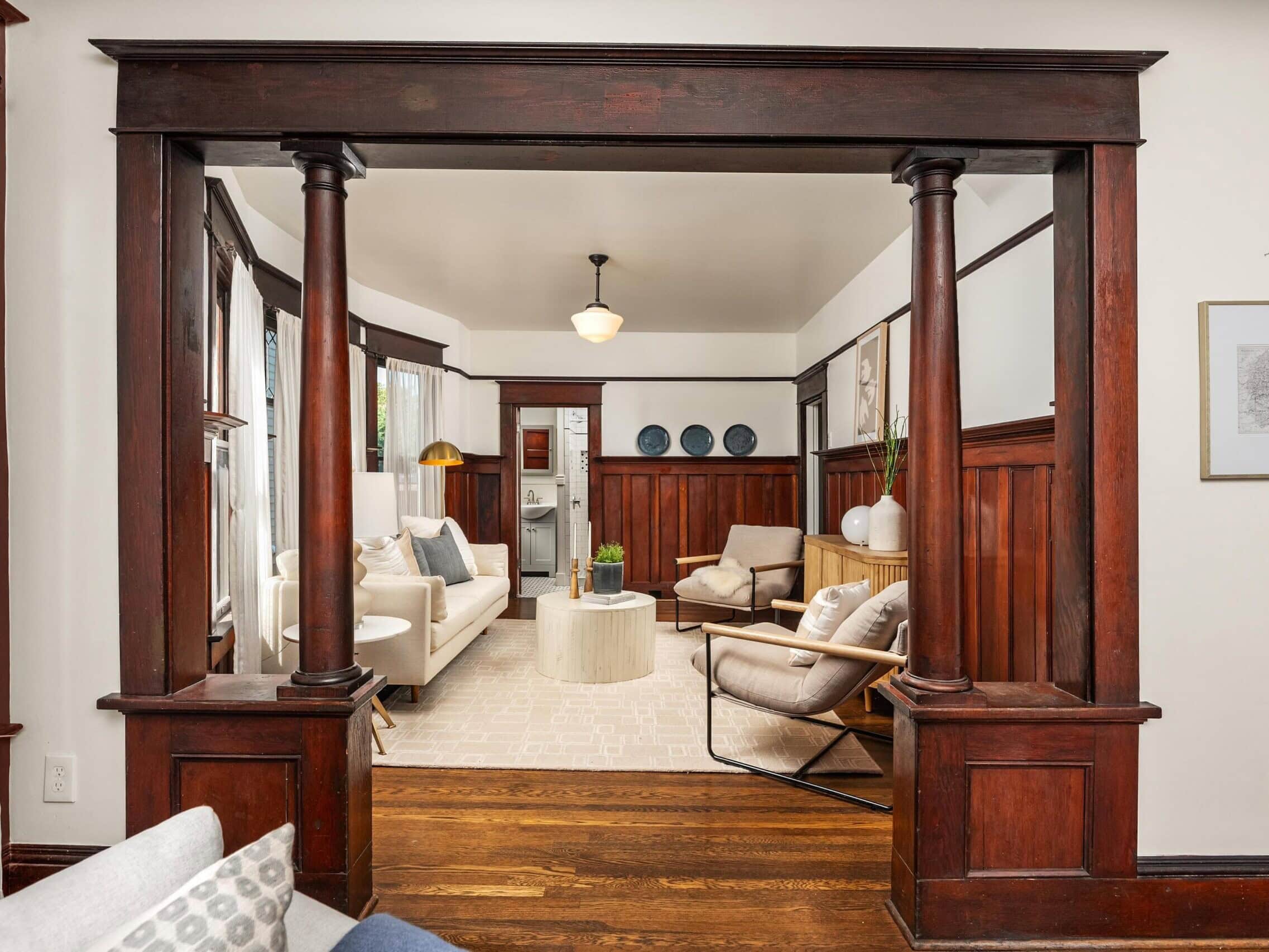 View of a cozy living room with wooden columns and trim, neutral furniture, a round coffee table, artwork on the walls, and light streaming in from multiple windows.