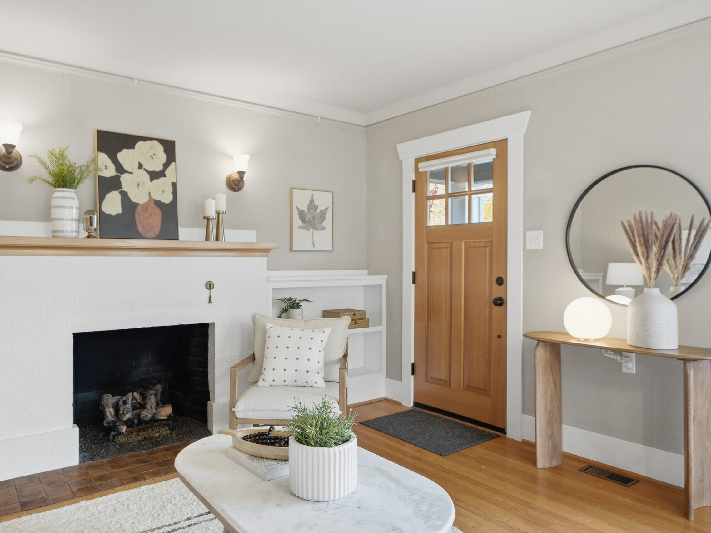 A cozy living room with a white fireplace, wooden mantel, beige walls, and a wooden front door. A round mirror and lamp sit on a small table near the door. Plants and art decorate the space.