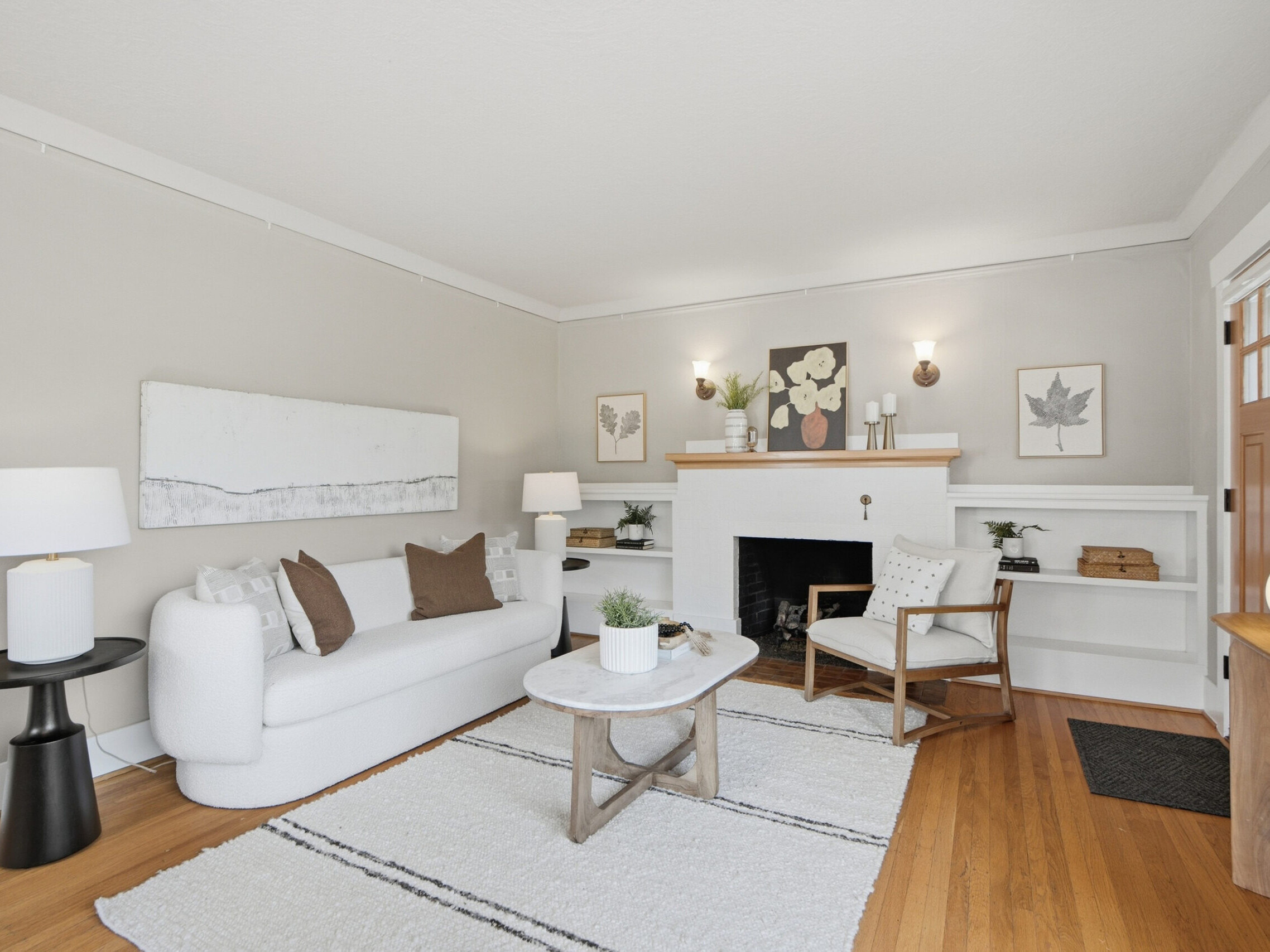 Bright living room with white walls, a white sofa, wooden chair, fireplace, built-in shelves, and light wood floors. Neutral decor includes artwork, lamps, and plants, creating a cozy and inviting atmosphere.