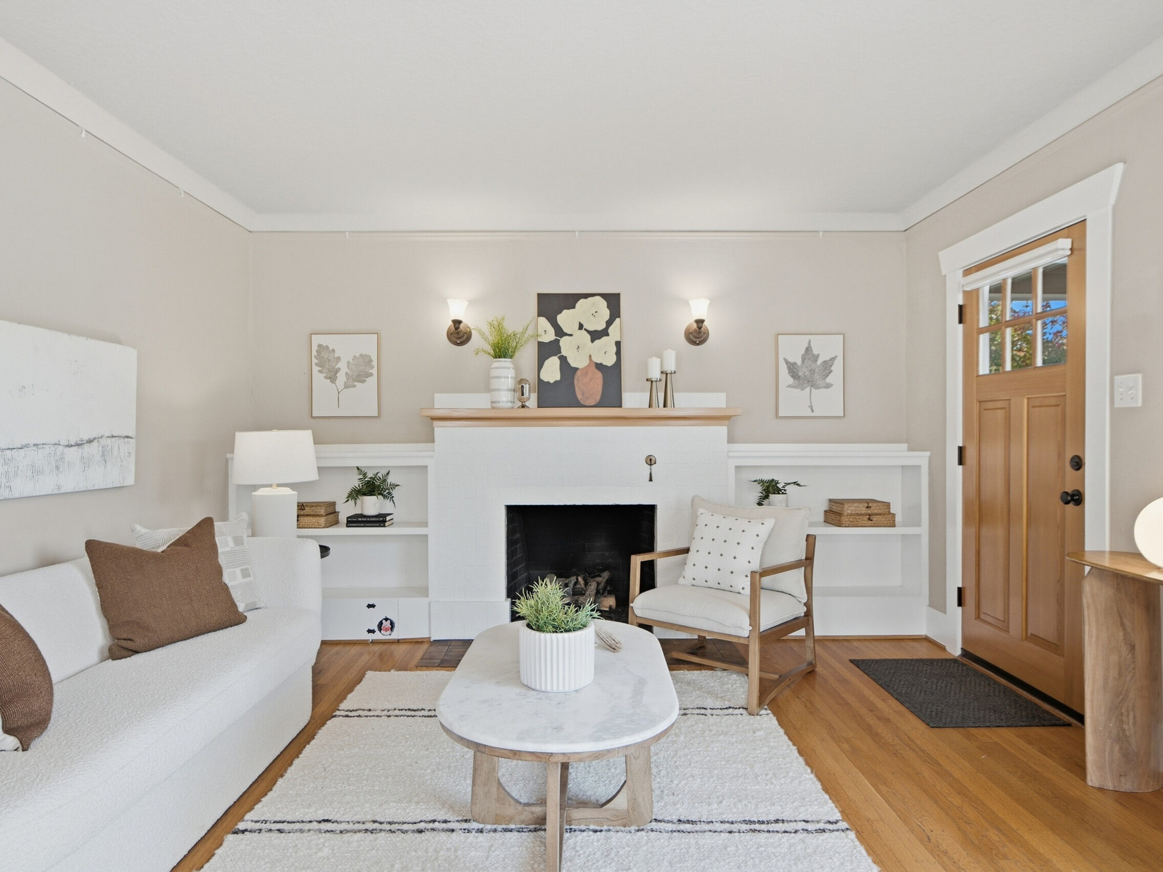 Bright living room with neutral decor, white sofa, wooden armchair, coffee table, fireplace with art above, built-in shelves, and wooden front door. Light walls, hardwood floor, and minimalist accessories create a cozy, modern feel.