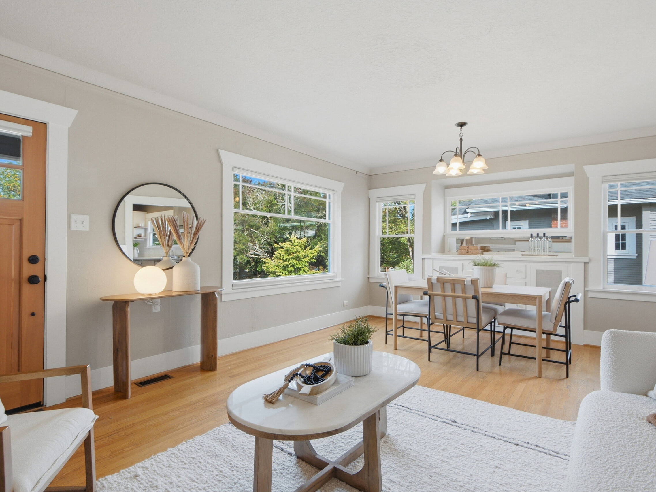 Bright, modern living and dining area with light wood floors, beige walls, large windows, a round mirror, a wooden table, a white sofa, and a dining table with chairs near the windows.