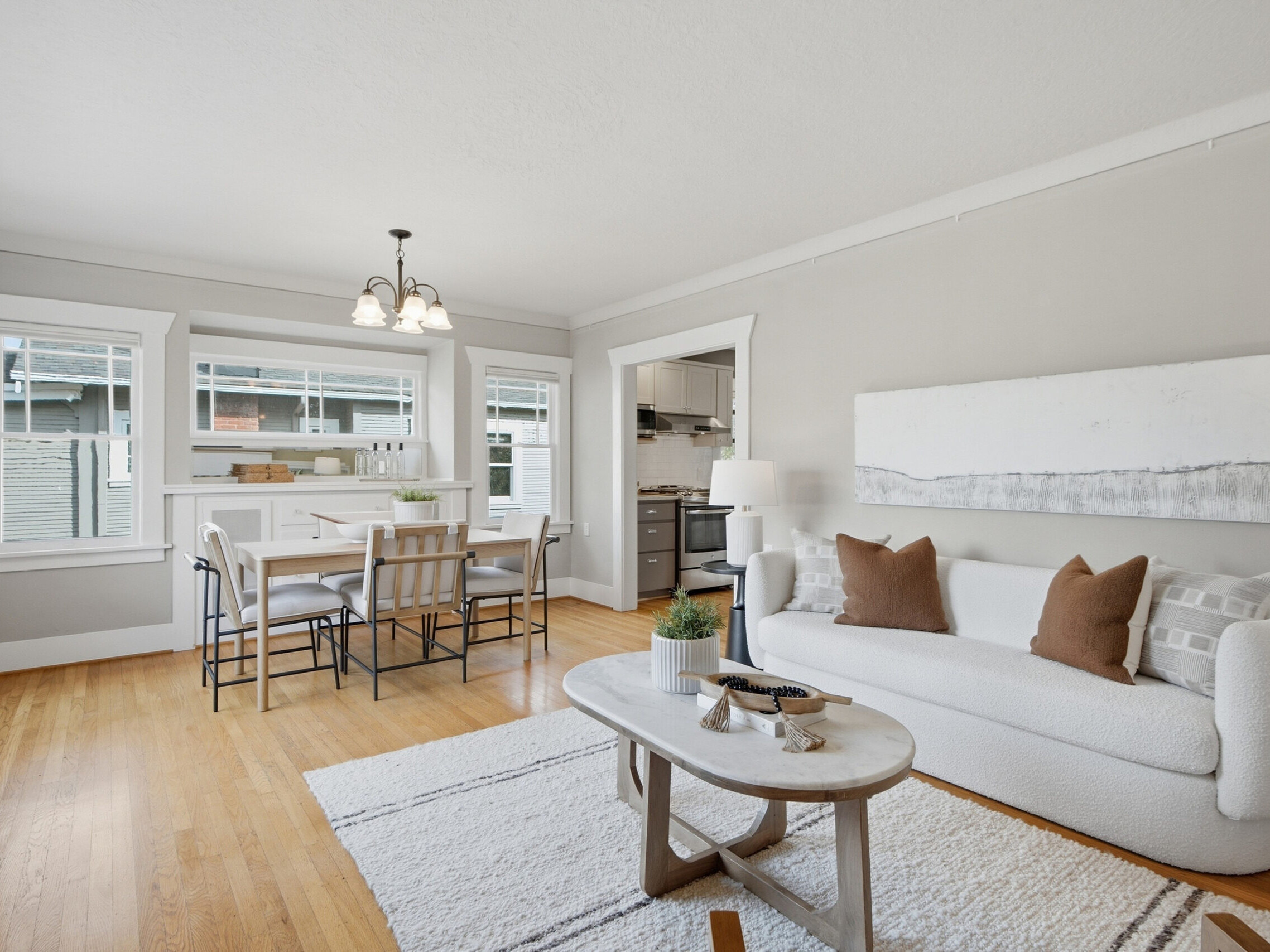 Bright living and dining area with light walls, wood flooring, a white sofa with brown pillows, a light rug, and a dining table with four chairs near windows. Kitchen is visible through an open doorway.