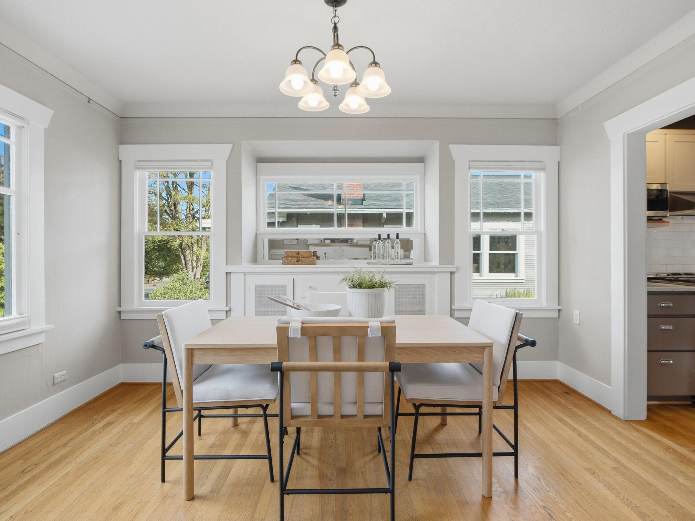 Bright dining room with a wooden table, four chairs, hardwood floors, large windows, built-in shelves, and a hanging light fixture. White walls and trim create a clean, modern look. Kitchen visible to the right.