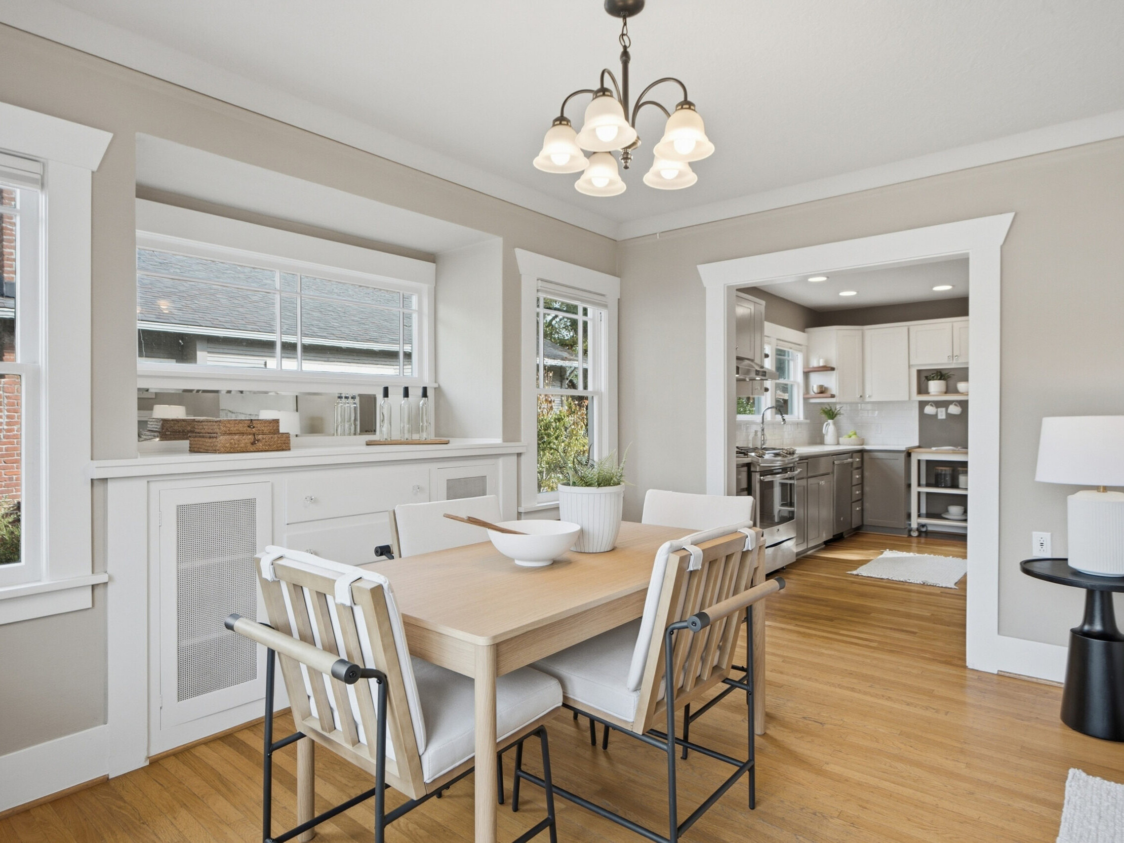 A bright dining room with a wooden table, four chairs, light fixture, built-in cabinetry, hardwood floors, and view into a modern kitchen. Neutral decor and natural light create a welcoming atmosphere.