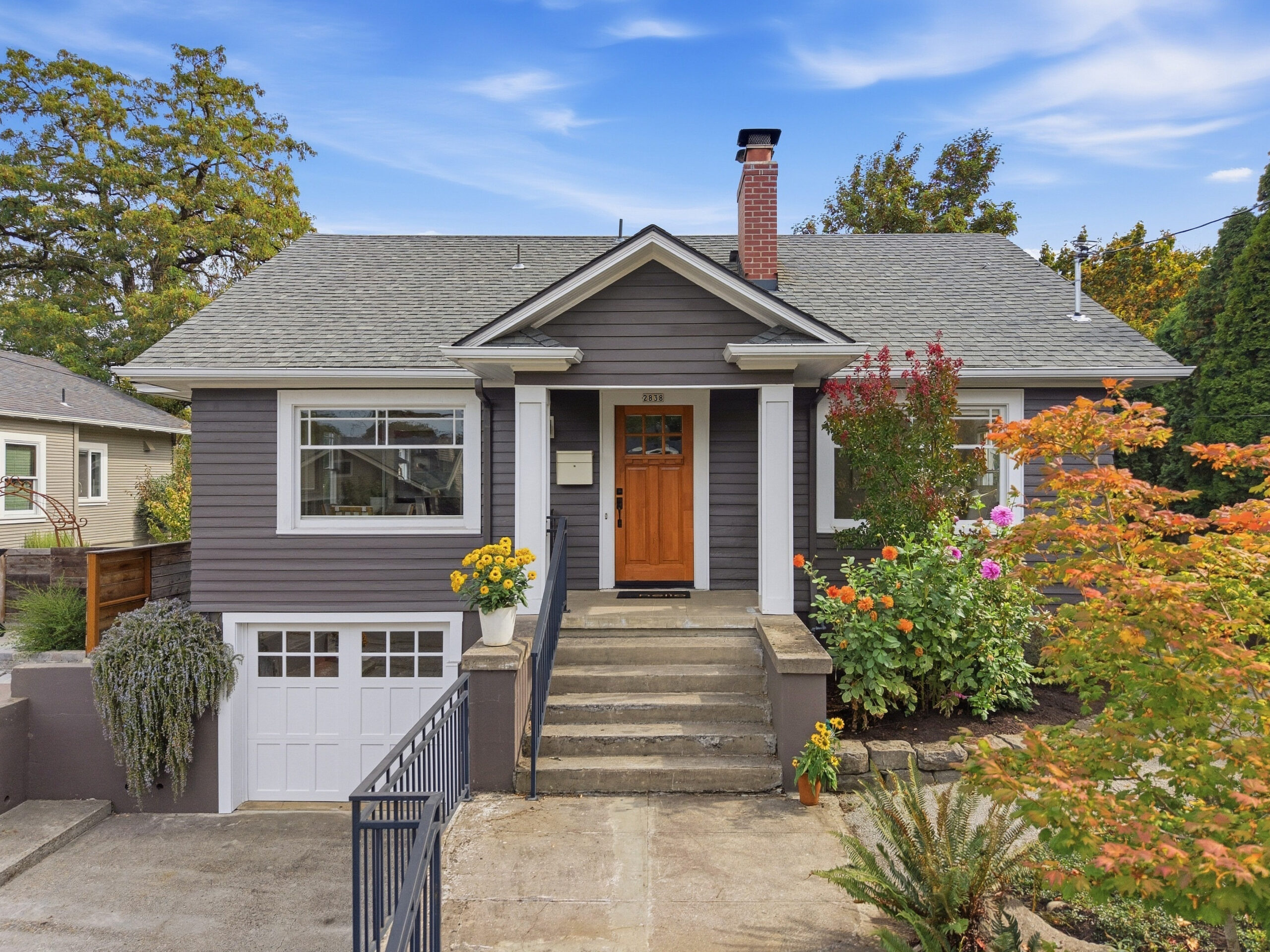 Gray house with white trim, wooden front door, small front porch, steps leading up, garage below, colorful flowers in pots and garden beds, trees with green and orange leaves, blue sky with clouds.