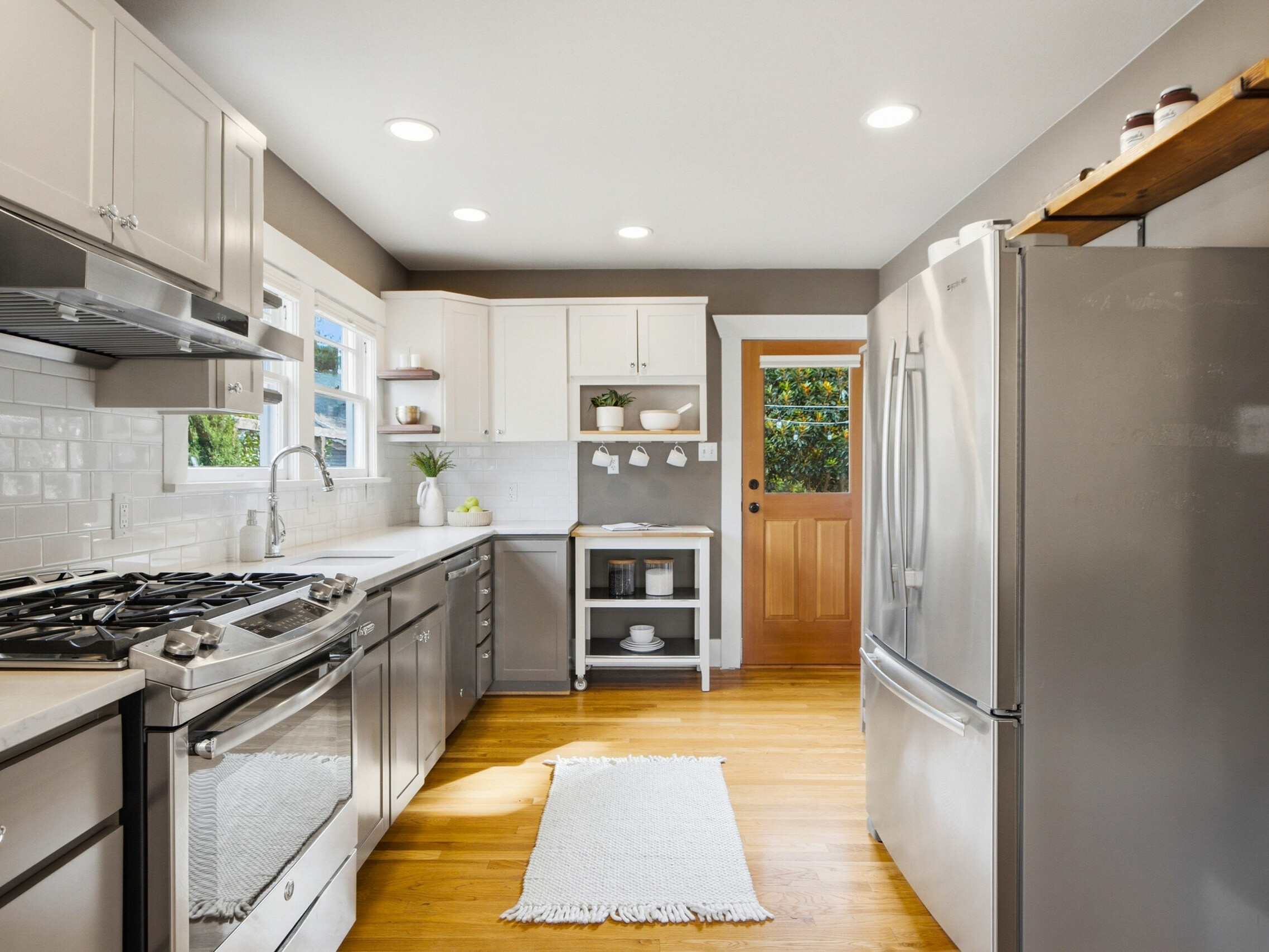 Modern kitchen with stainless steel appliances, white cabinets, a gas stove, subway tile backsplash, wooden floors, a small white rug, and a wooden door with glass, with natural light streaming in.