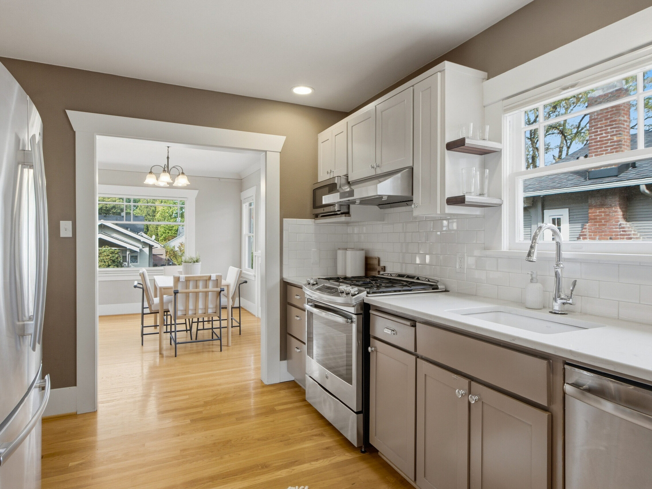Modern kitchen with white cabinets, stainless steel appliances, and wood flooring leads to a bright dining area with a window and a light fixture above a table with chairs.