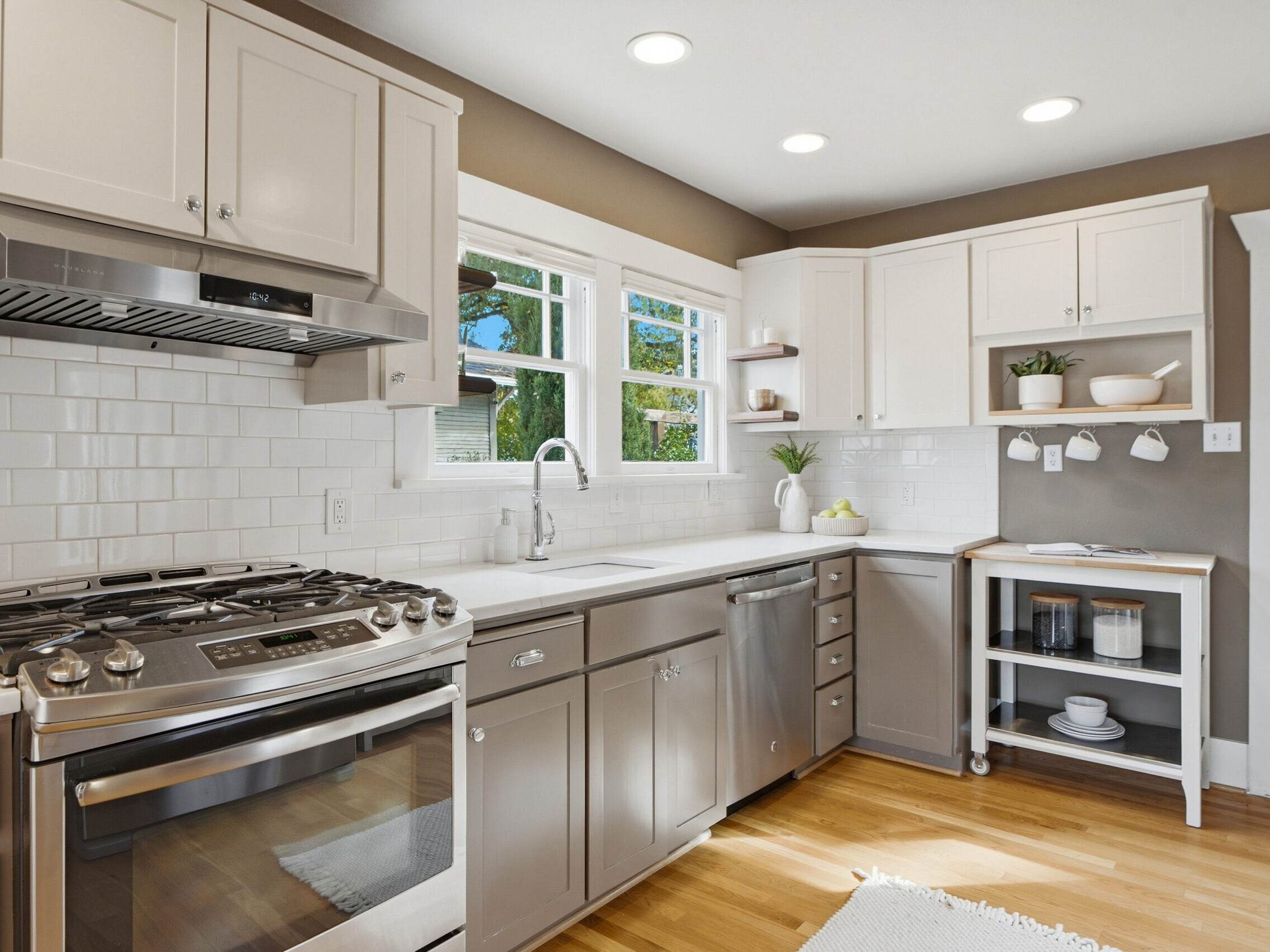 Bright kitchen with white cabinets, stainless steel appliances, subway tile backsplash, wood floors, a window over the sink, and a small corner shelf holding dishes and a plant.