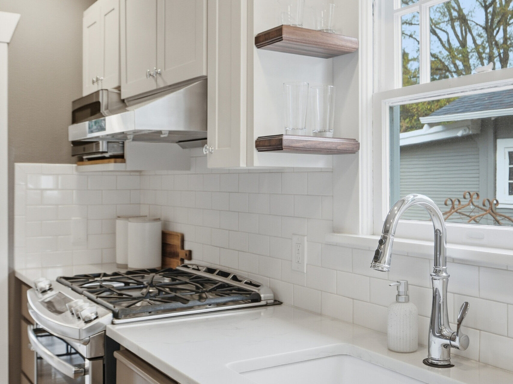 Modern kitchen with white cabinets, gas stove, white subway tile backsplash, stainless steel oven, floating shelves with glasses, large window, and a white sink with a sleek faucet.