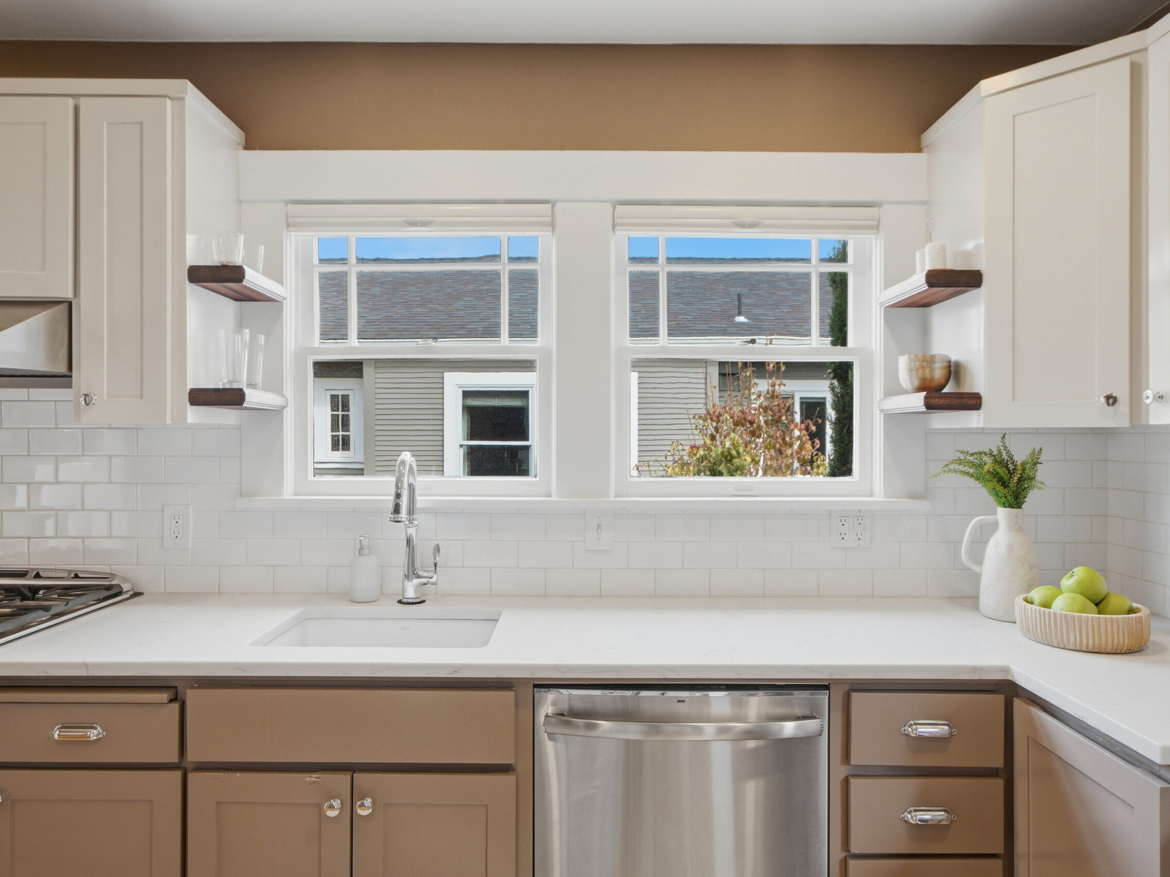 Modern kitchen with brown and white cabinets, white countertop, stainless steel dishwasher, sink under two large windows, and a bowl of green apples on the counter.