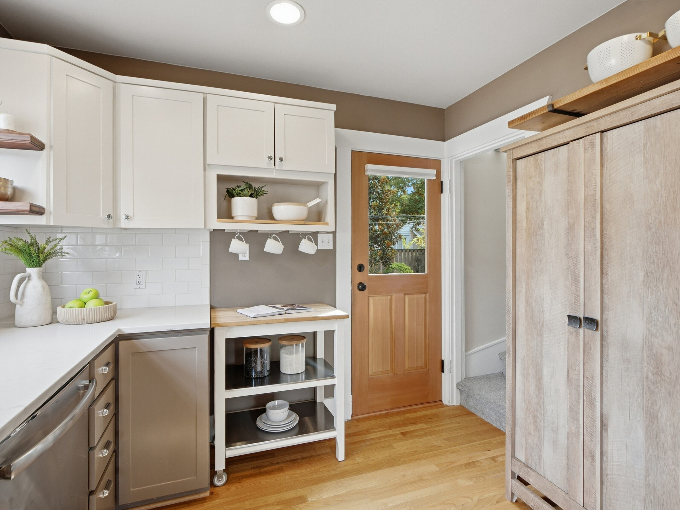 Bright kitchen with white cabinets, wood accents, stainless steel dishwasher, and a small island cart. A wooden door with a window leads outside. Shelves and countertops display dishes, plants, and fruit.