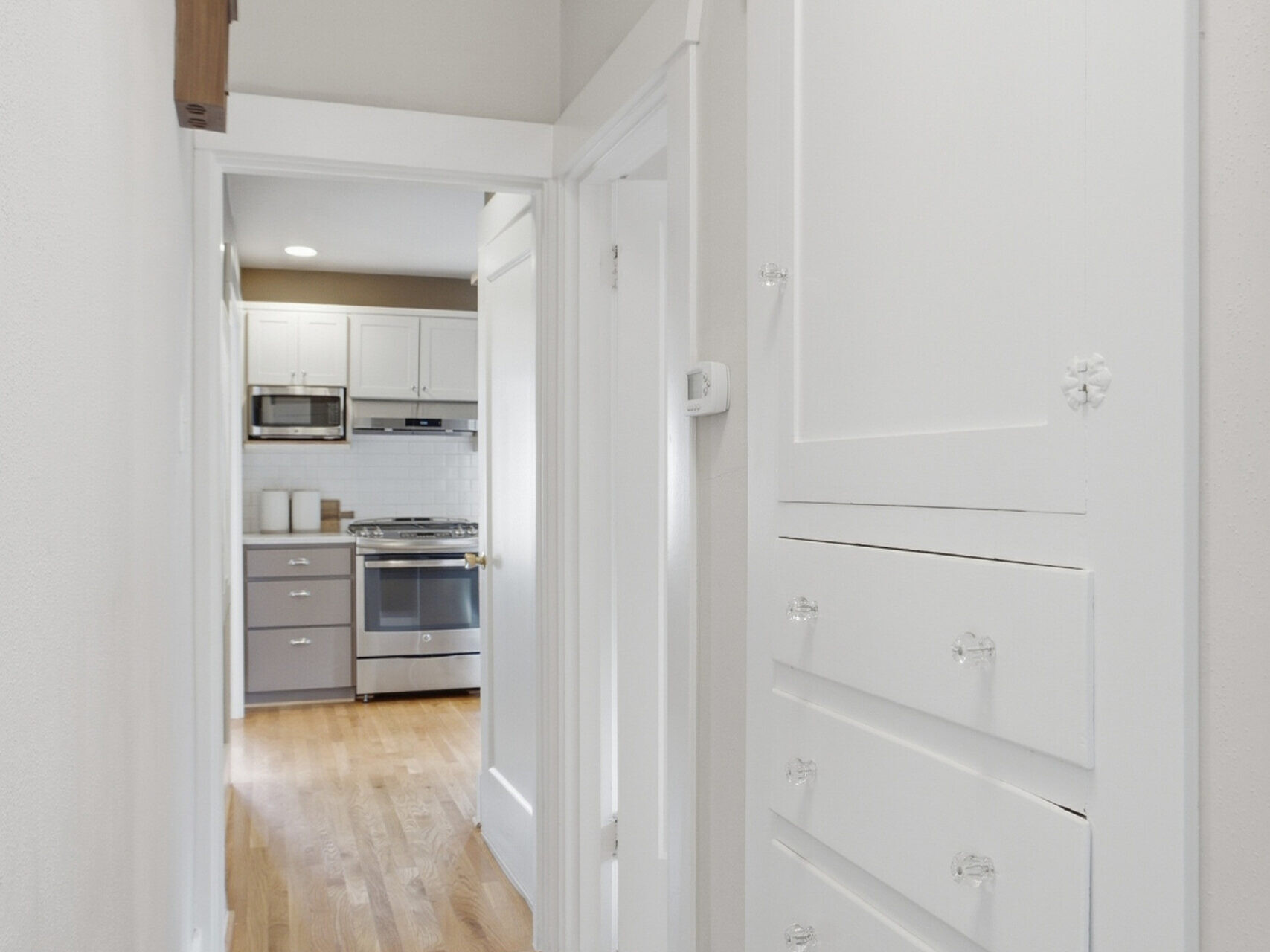 A narrow hallway with hardwood floors, built-in white cabinets on the right, and a view into a modern kitchen with stainless steel appliances at the end.